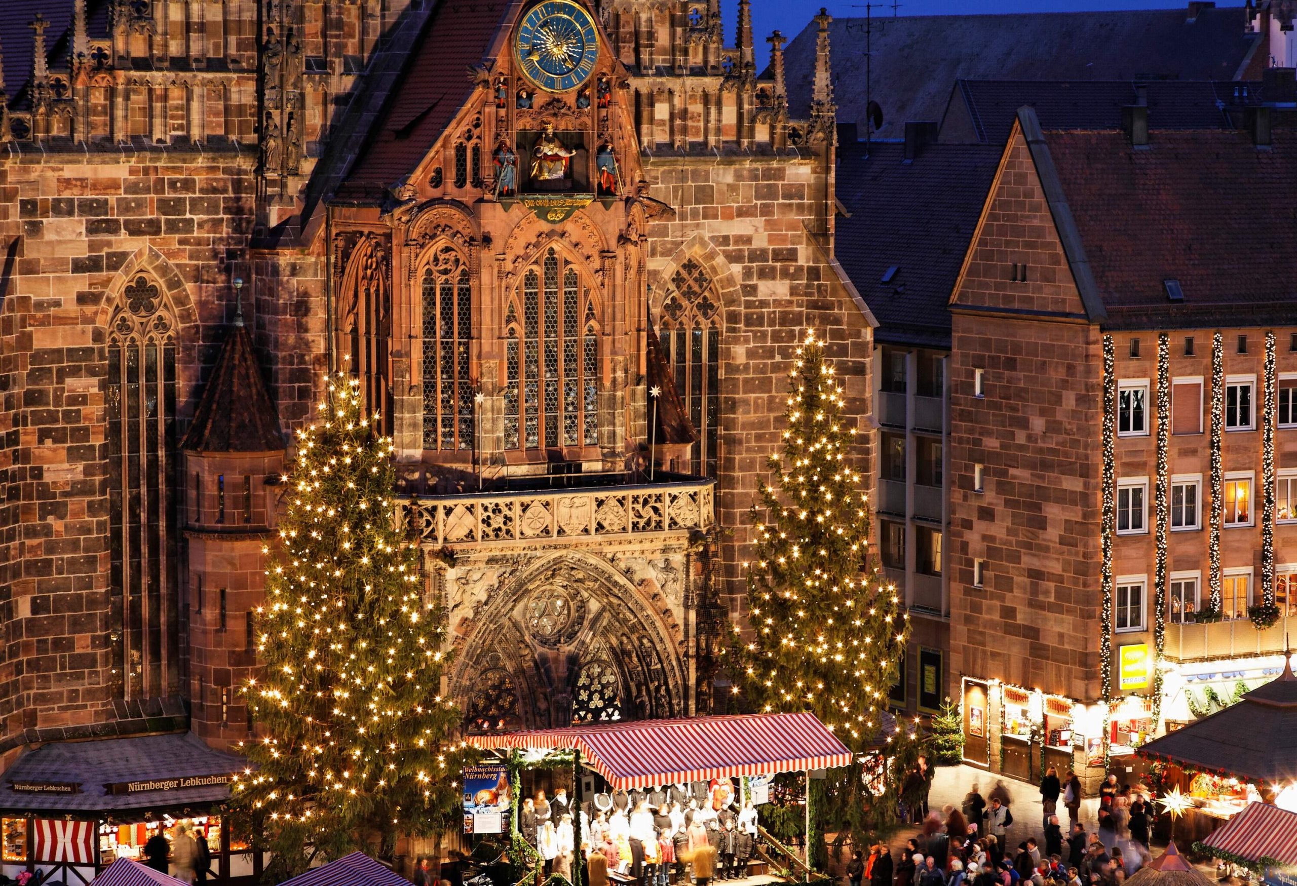 An elaborately decorated brick cathedral in Gothic style with tall Christmas trees on either side of its entryway and Christmas shops on the main square.