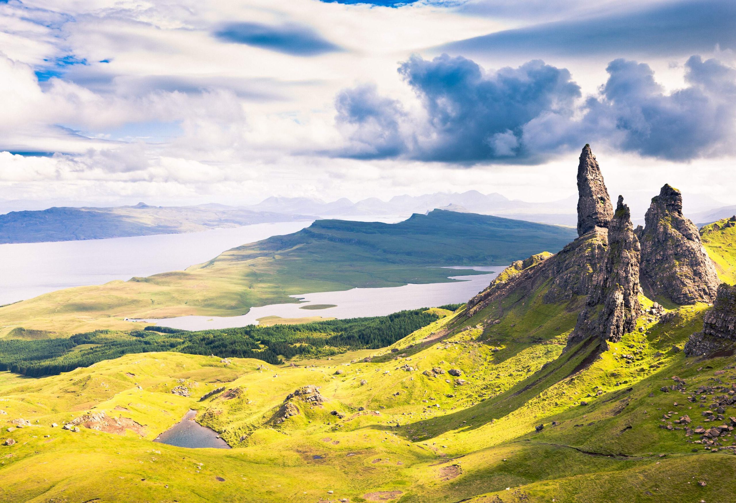 A lush mountainous terrain with three pointed rock formations overlooking a lake in the distance.