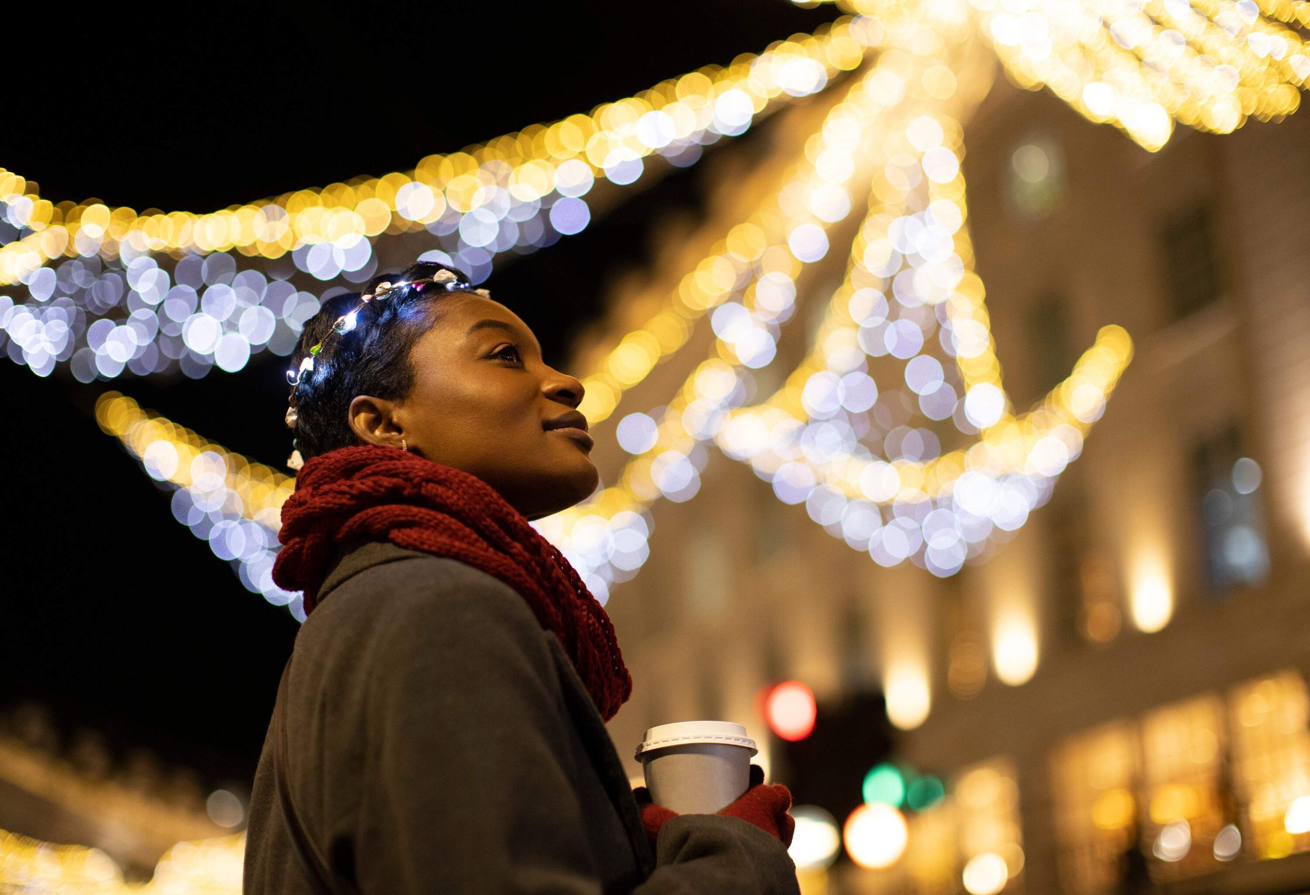 Carefree young woman in lit Christmas headband drinking coffee in city with lights at night