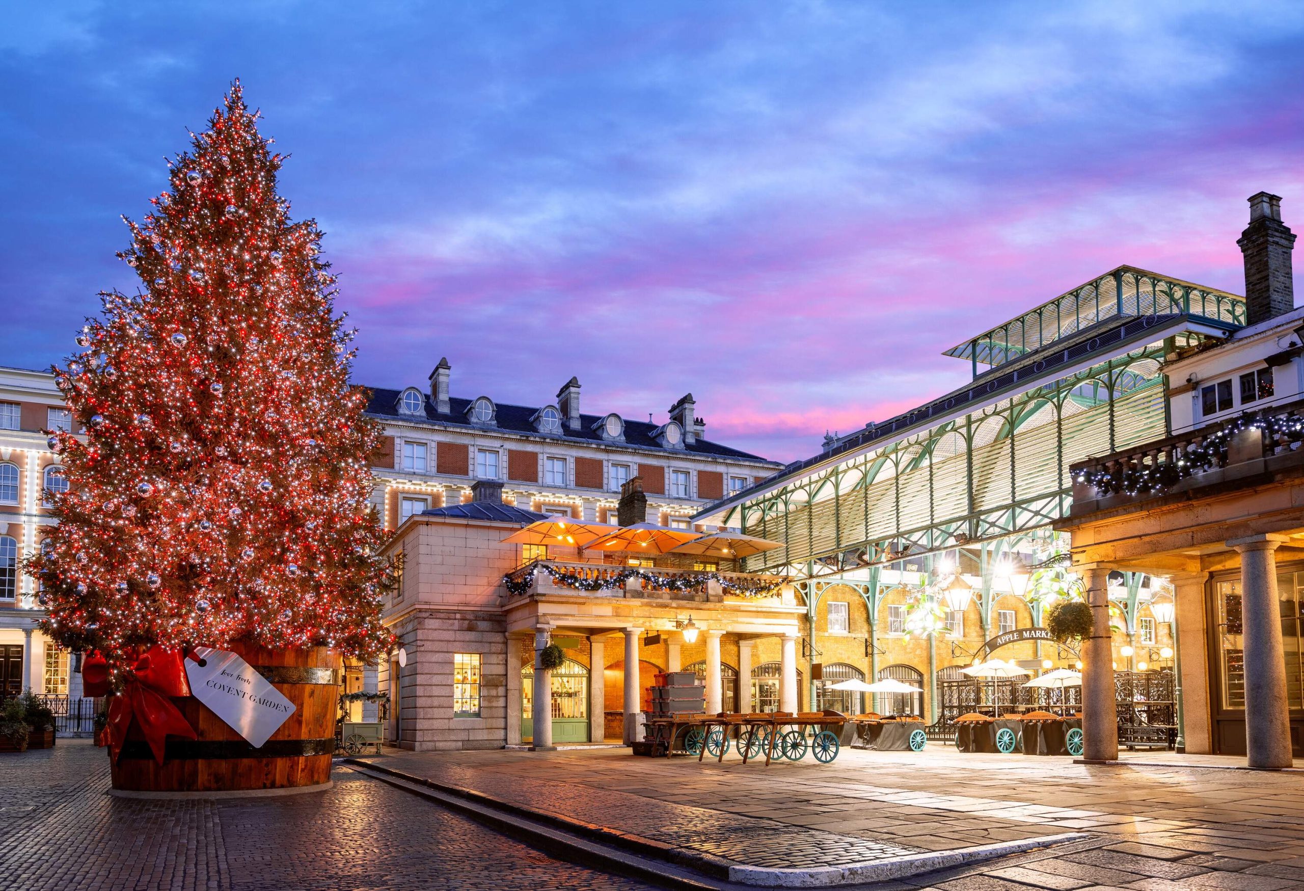 A giant illuminated Christmas tree on a bucket with a red ribbon in front of the brightly lit covered market.