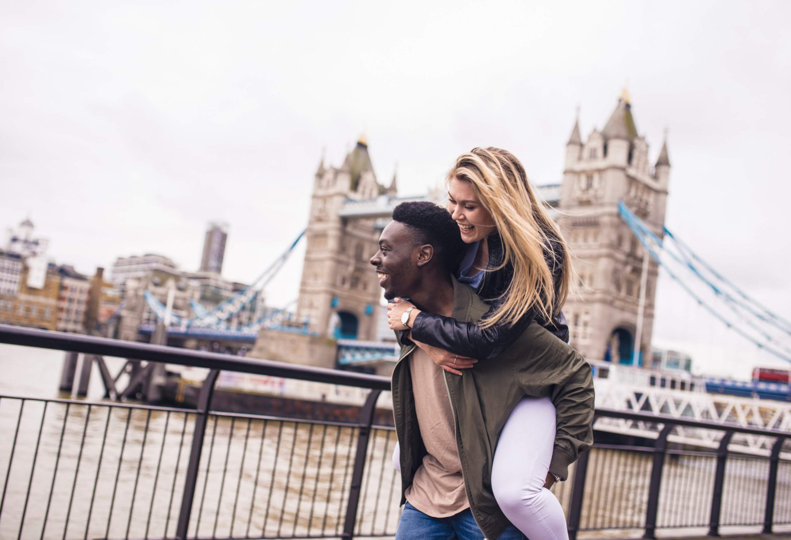 An out-of-focus river and the Tower Bridge form the background for a happy couple, with the woman riding on the man's back.