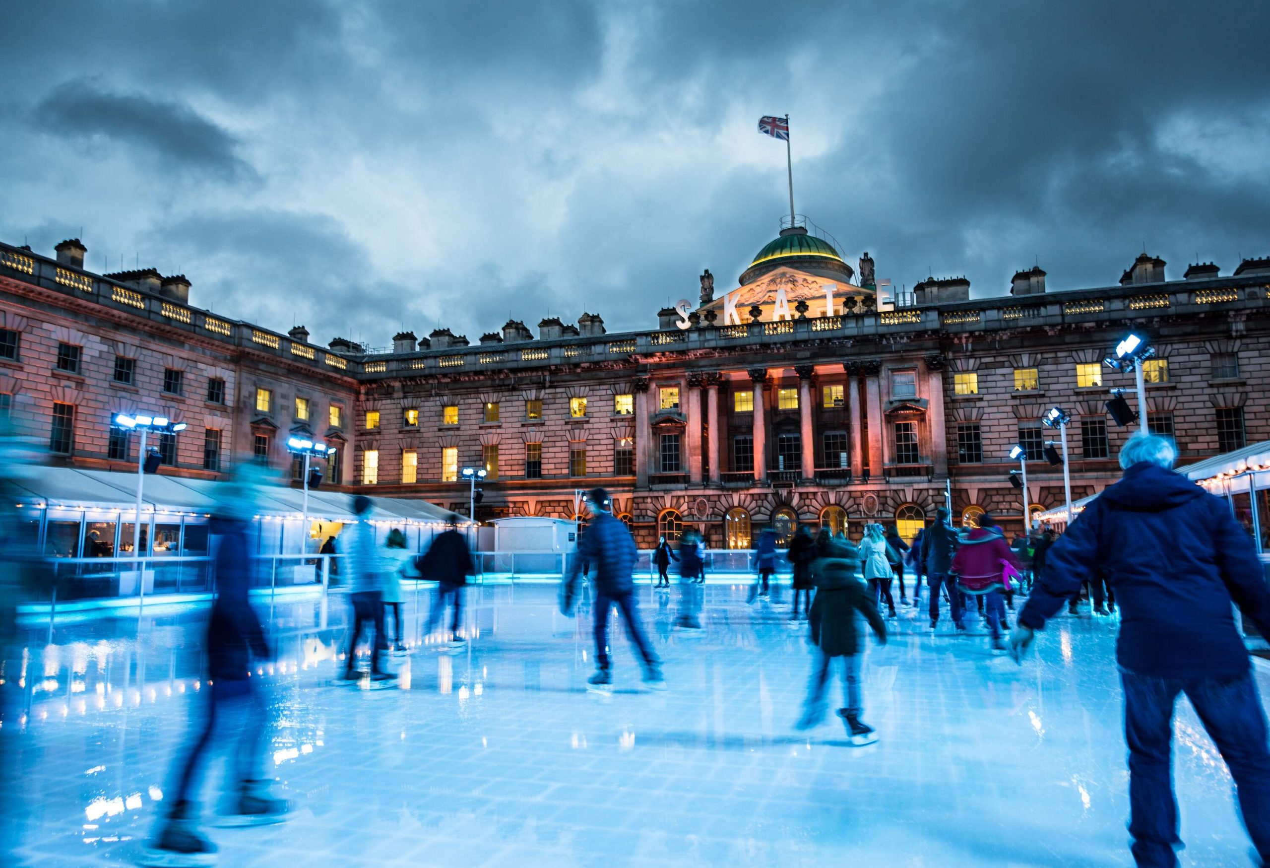 Blurred motion of crowds of people ice skating at Somerset House, a publically owned building in central London. The image features warm evening light, and an ominous, moody sky. The people appear as unrecognisable blurs due to the long exposure used. Somerset House is a large Neoclassical building situated on the south side of the Strand in central London, overlooking the River Thames, just east of Waterloo Bridge.