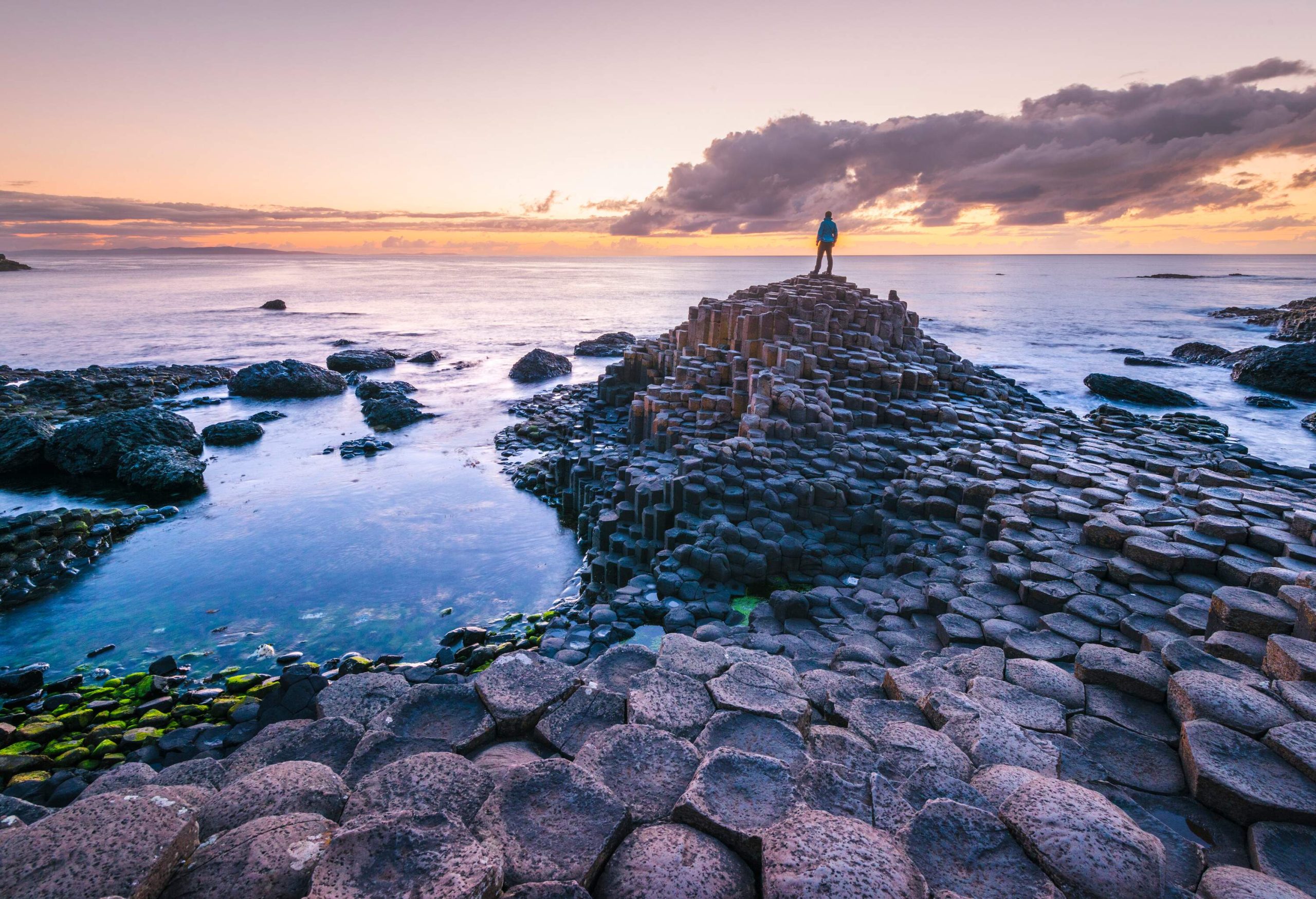 A person standing on the edge of an iconic landform featuring majestic basalt columns rising from the sea against the backdrop of a scenic twilight sky.