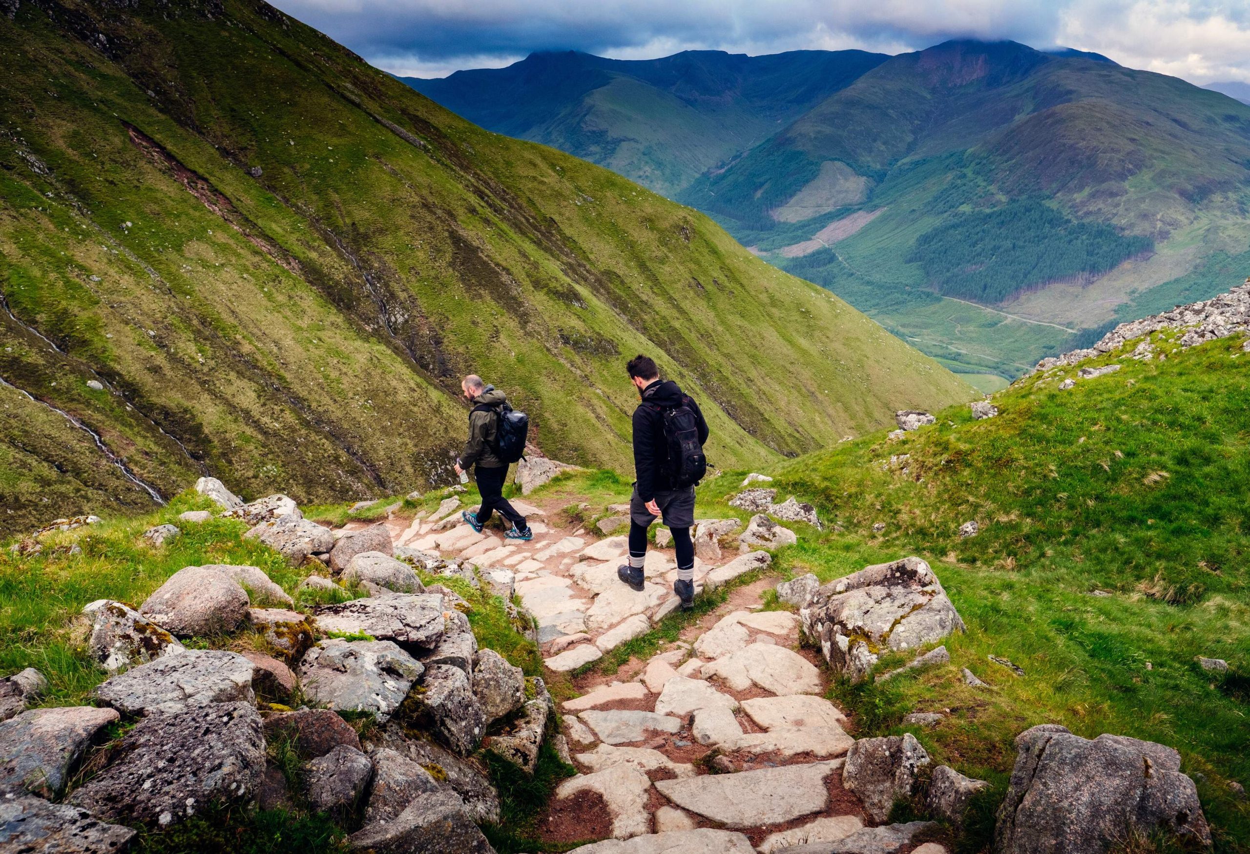 Two male hikers embark on an adventurous journey, descending a rugged stone path on the slope of a lush mountain.