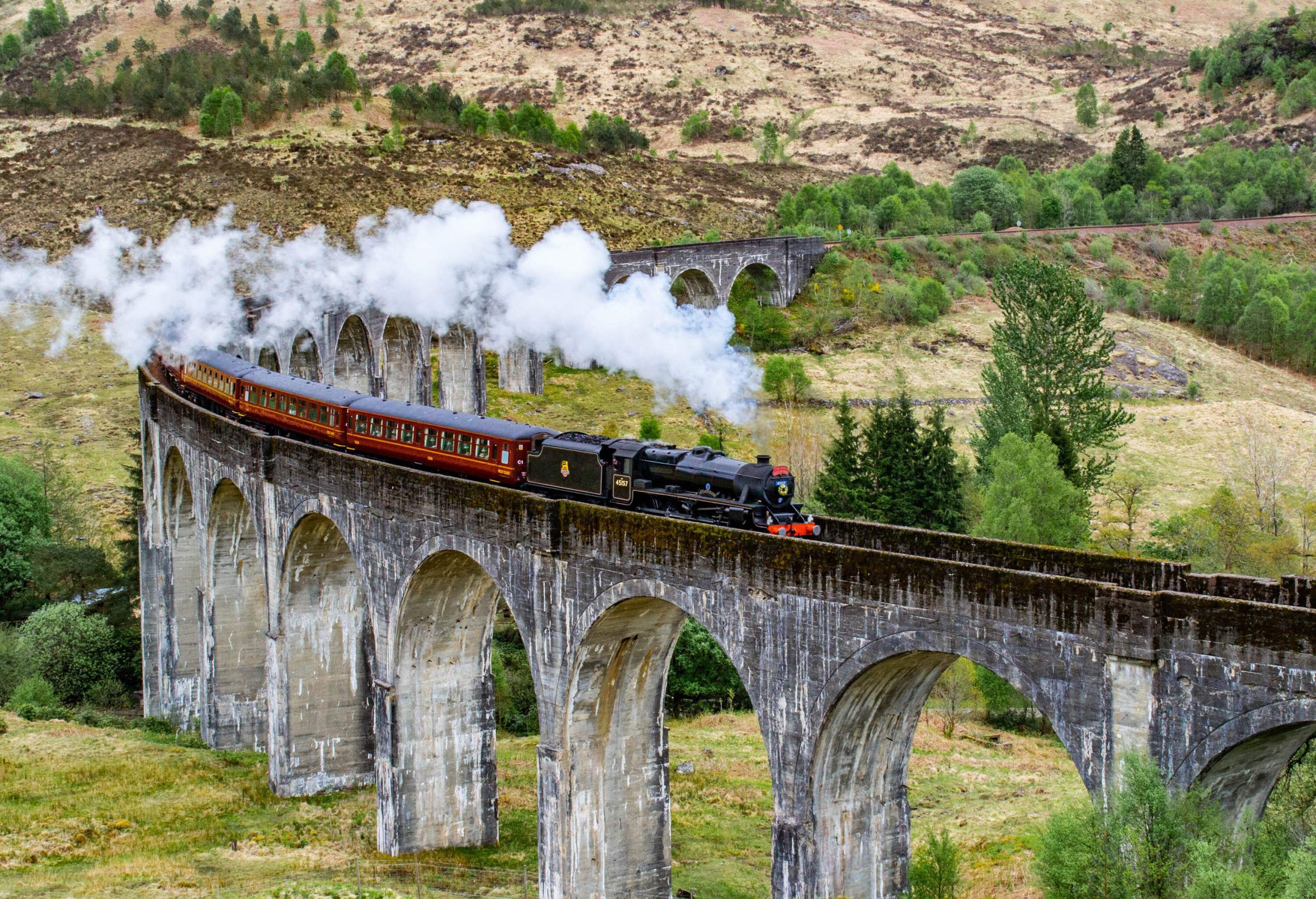 A steam train travelling on a long archway bridge in the mountains.