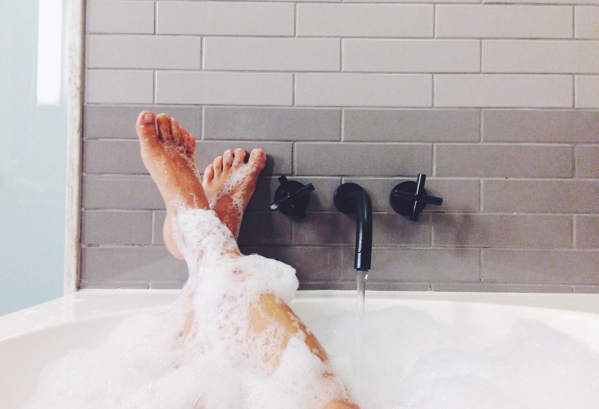 A person's legs, covered in soapy suds, are raised and resting on the edge of a bathtub.