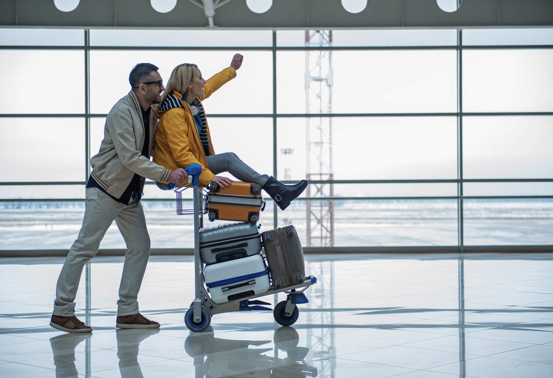 A happy blonde lady sits on a trolley with a pile of luggage pushed by her lovely boyfriend in the terminal lounge.