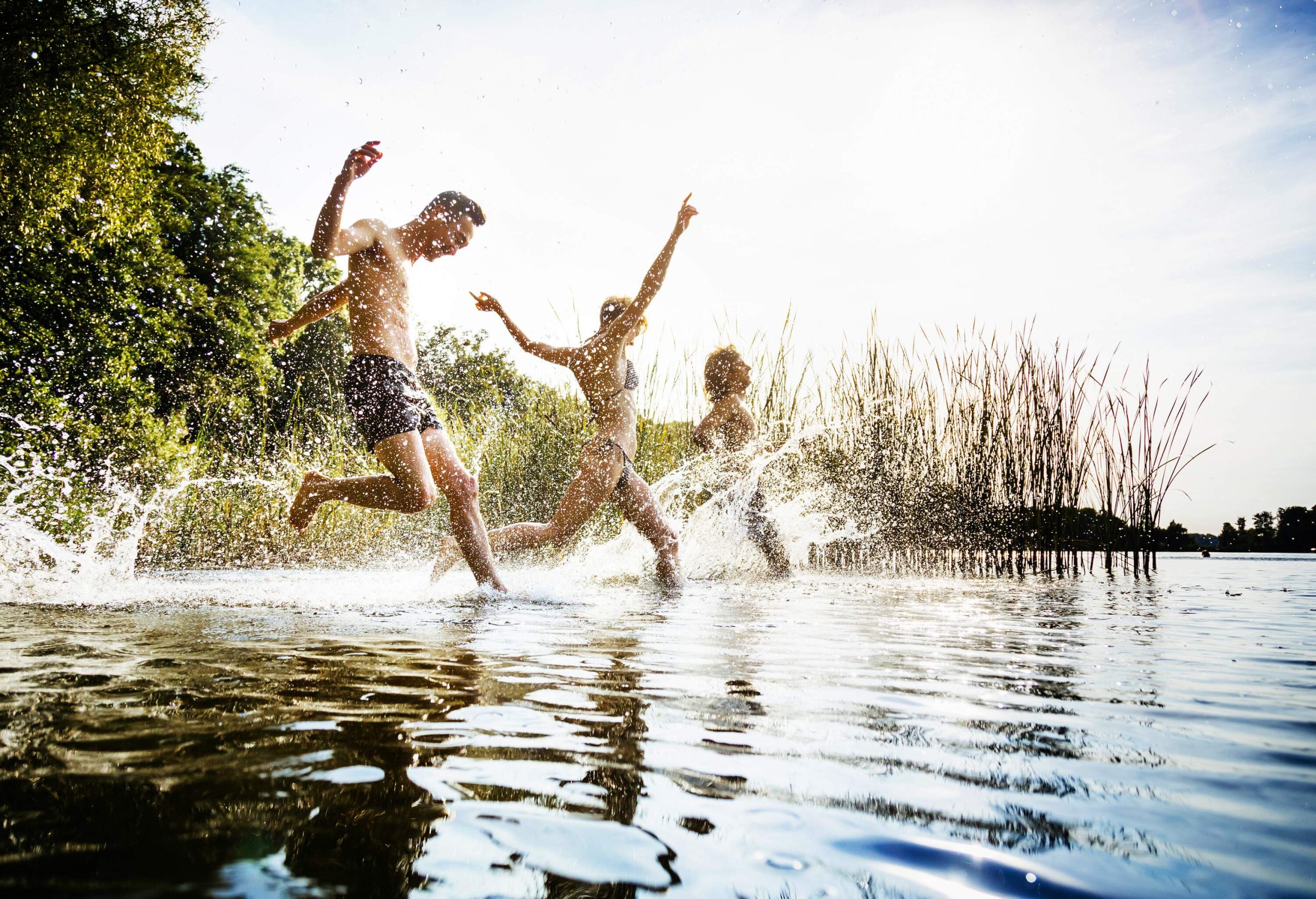 Three friends jumping into a lake and making water splashes as they go.
