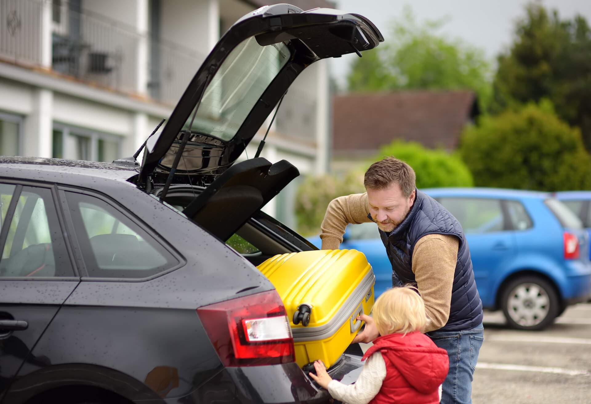 A man and a kid loading a yellow luggage into a black car's trunk.