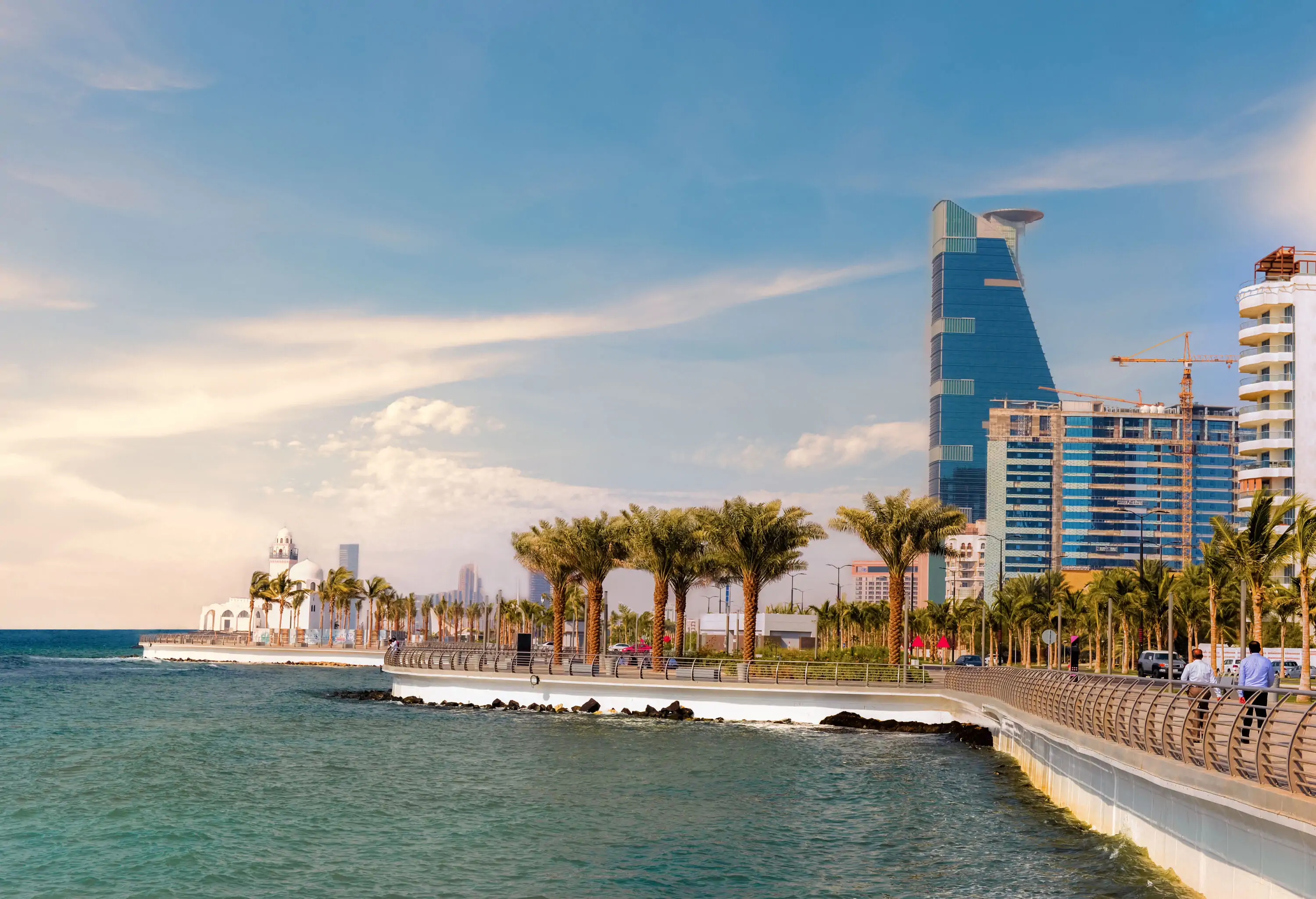 A picturesque promenade gracefully lines the coast, adorned with swaying palm trees, while towering buildings rise alongside.