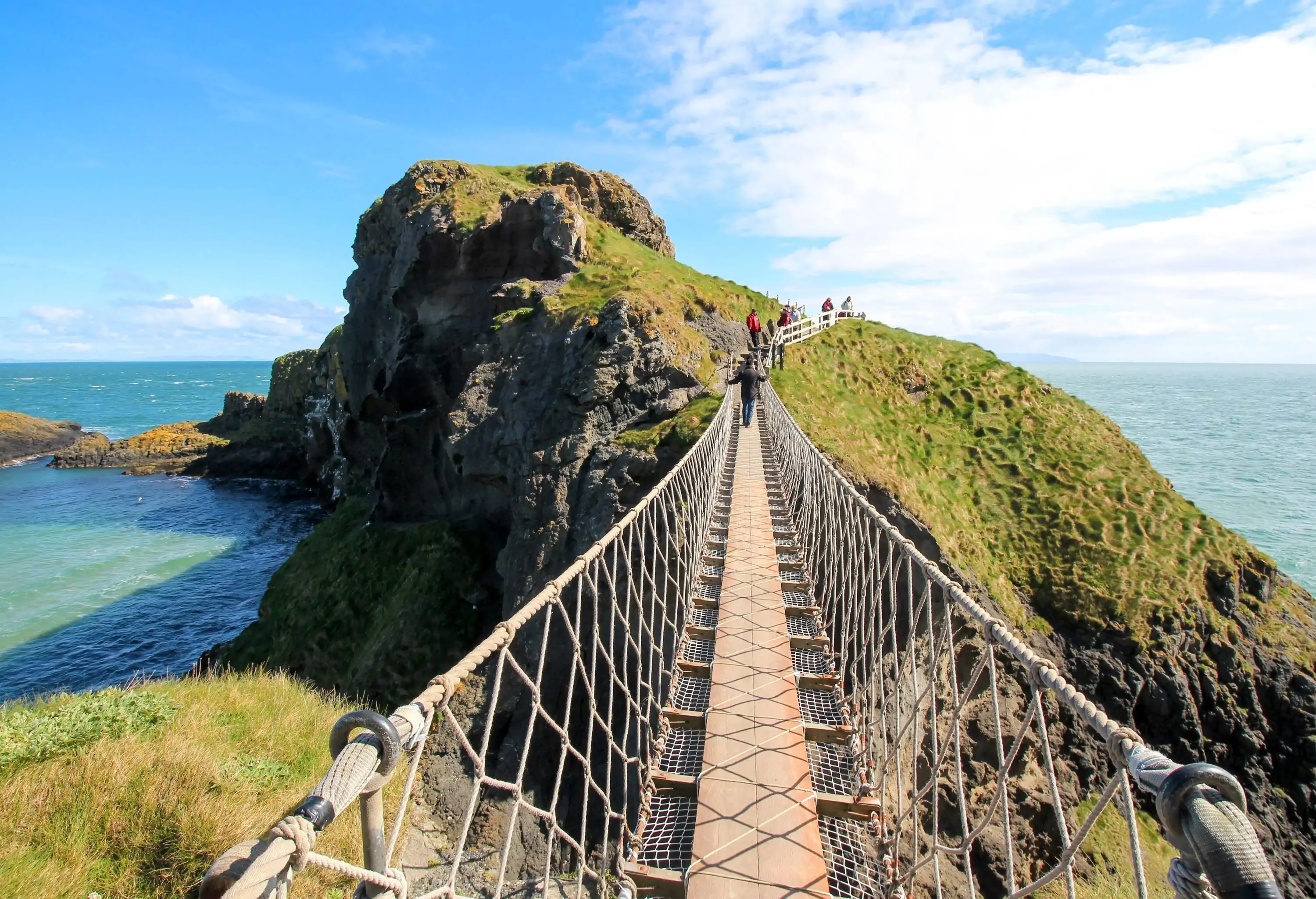A long, narrow rope bridge leads to lush, jagged cliffs overlooking the sea.
