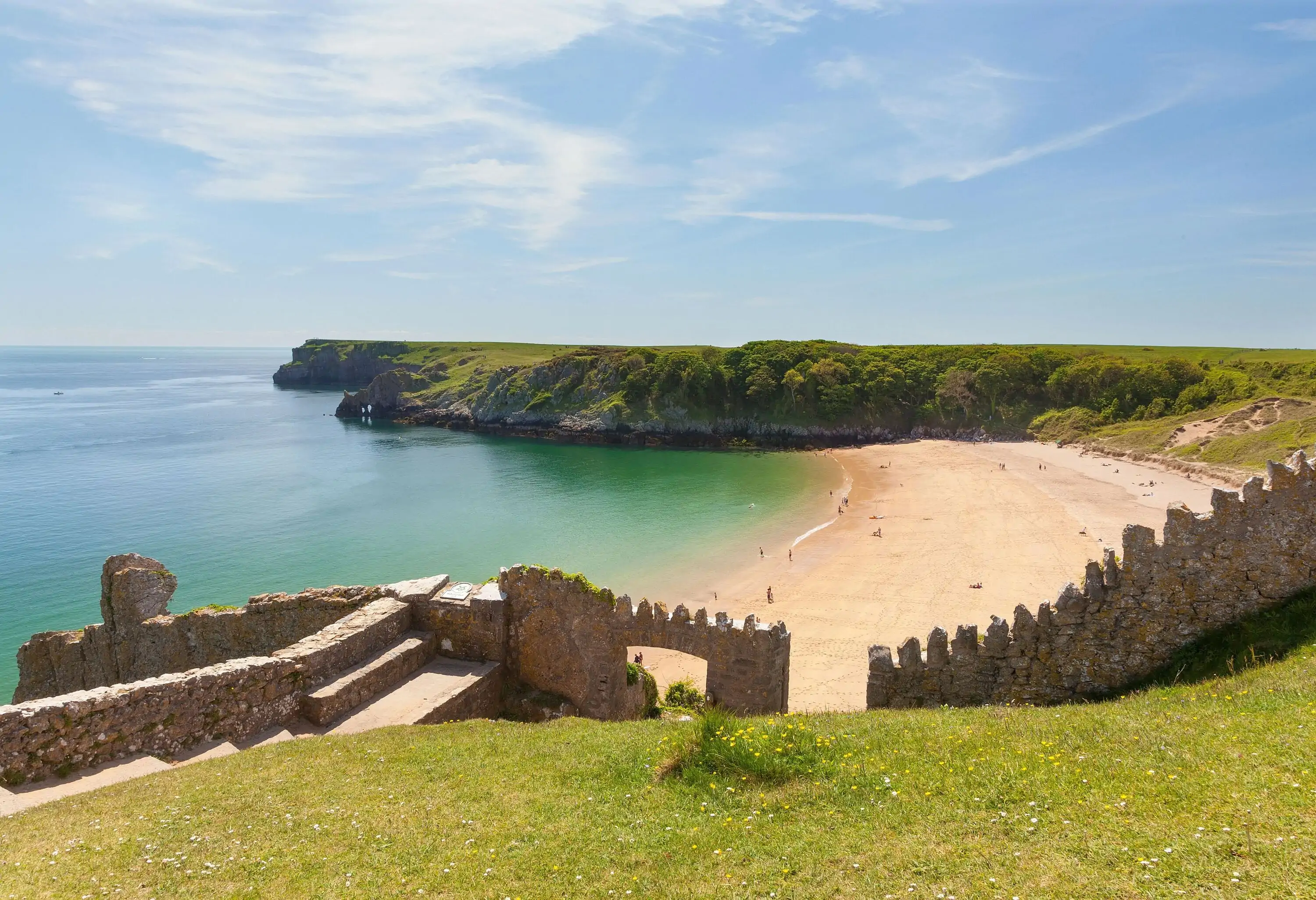 A ruined stone steps on a cliff leading to the sands of a quiet beach against a clear blue sky.