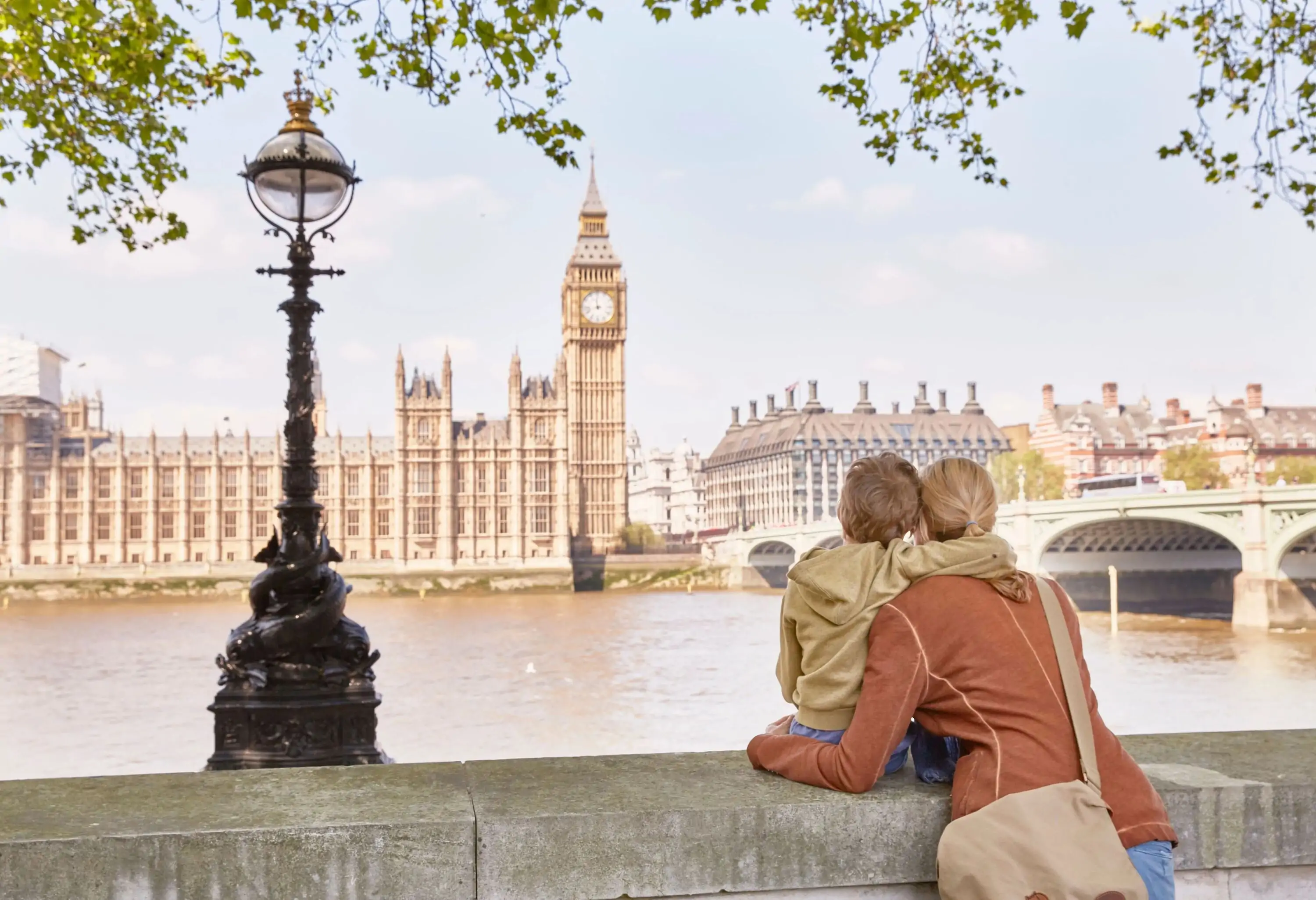 A child wraps his arms around the neck of a woman as they both look at the riverside Big Ben and Westminster Palace.