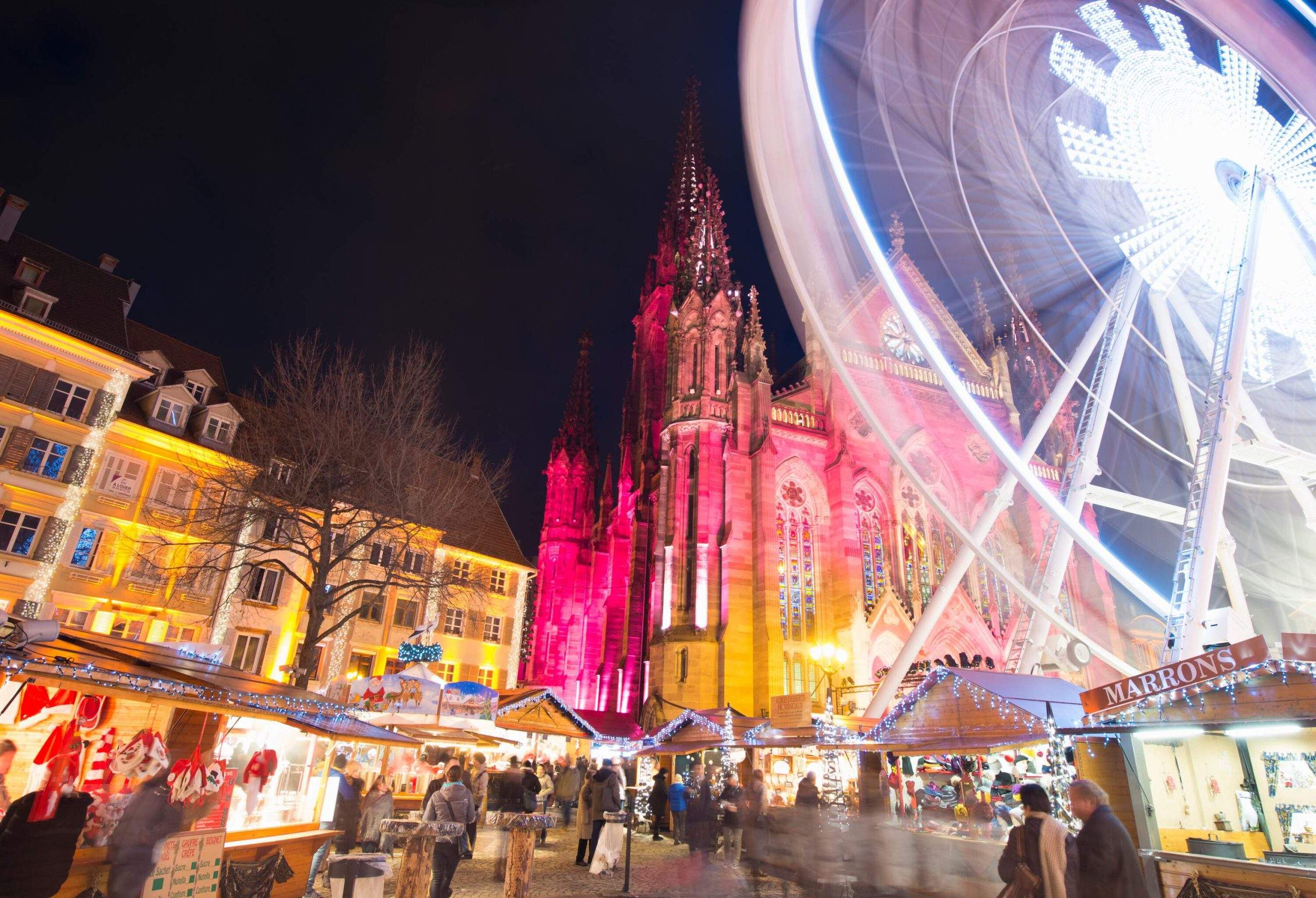A busy Christmas market with a Ferris wheel in front of a church lit up in a spectrum of colours.