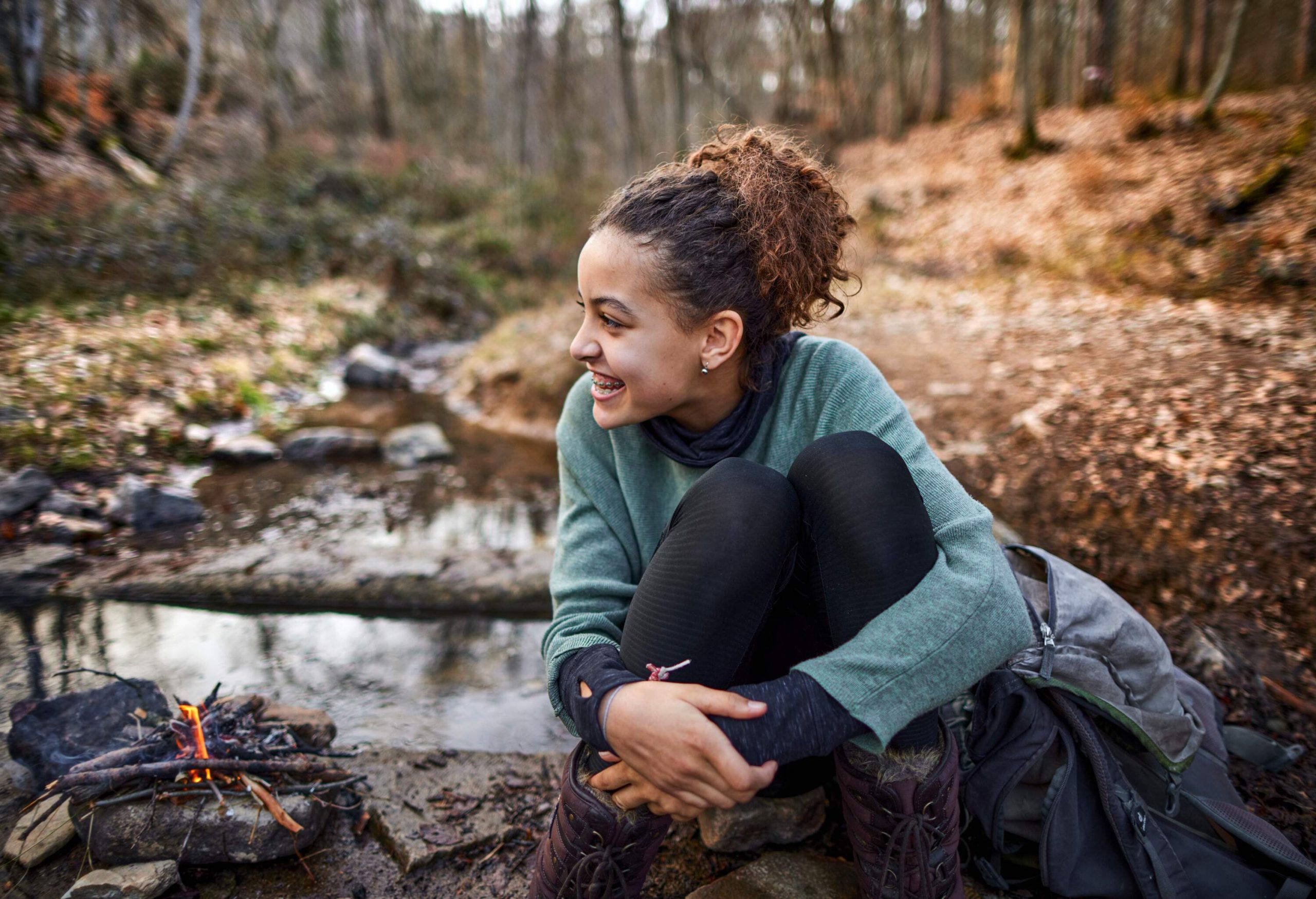 A young woman sitting on a rock next to a camp fire in the woods.