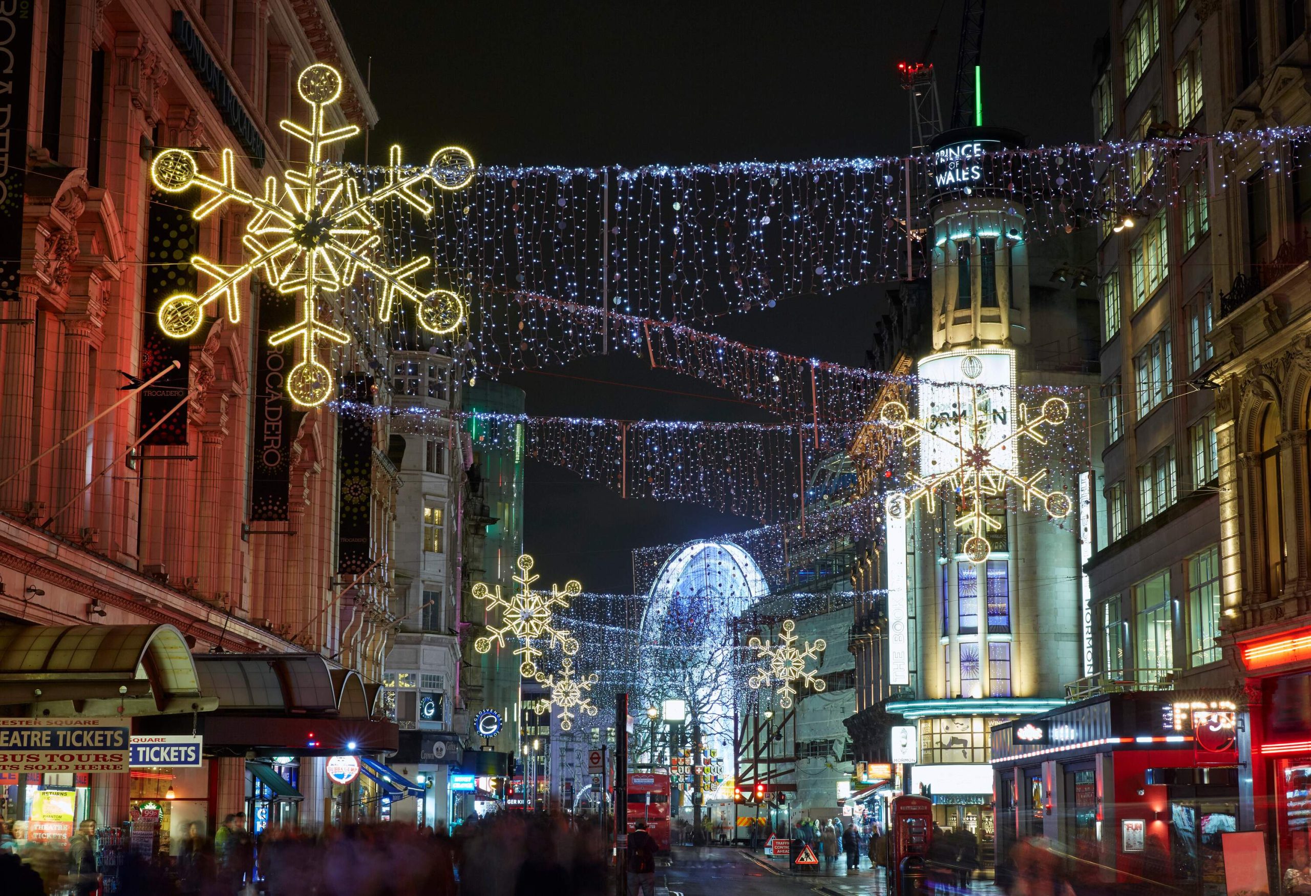 Brightly lit hanging Christmas lights in the centre of a commercial street full of people.