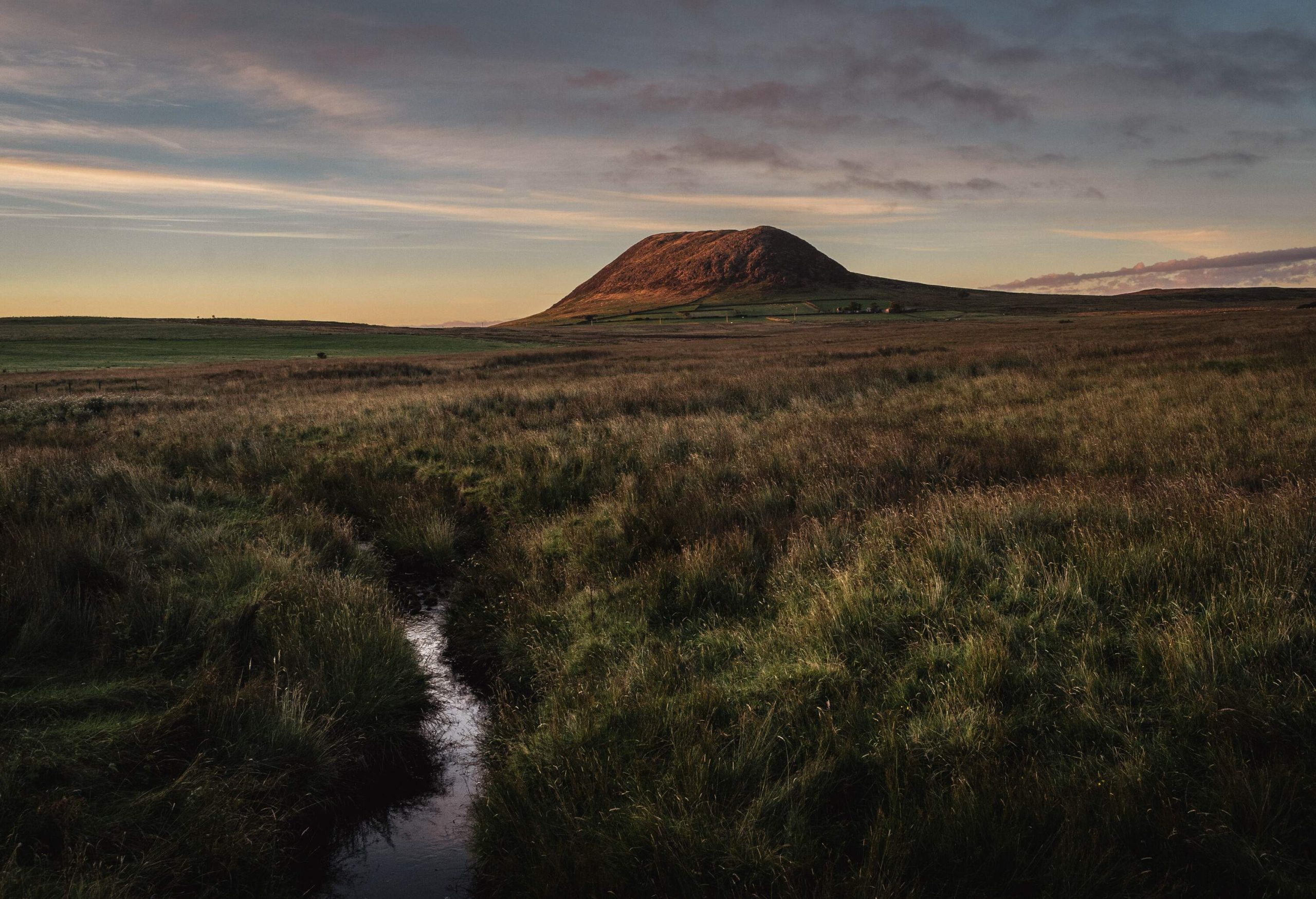 Slemish mountain on a summer evening at dusk in wide angle, with beautiful side lighting and overcast sky and a small water stream in foreground. The grass fields are bordered in distance with hand built stone walls. The mountain is a popular tourist attraction, and for walking. St. Patrick tended sheep there as a young man. The mountain is actually the plug of an extinct volcano.