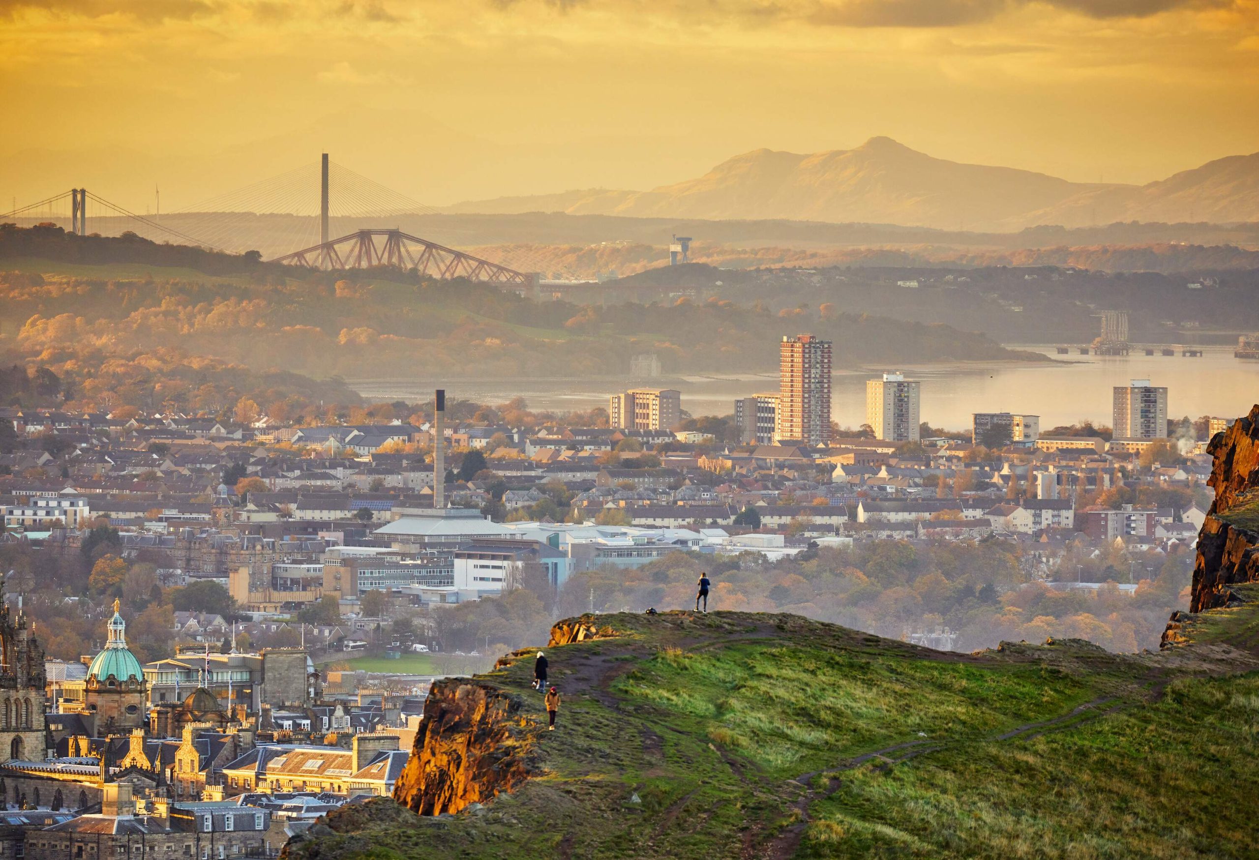 At the edge of town, three people stand on a cliff and stare out over the city and its many buildings and bridges, as well as the surrounding hills and mountains.