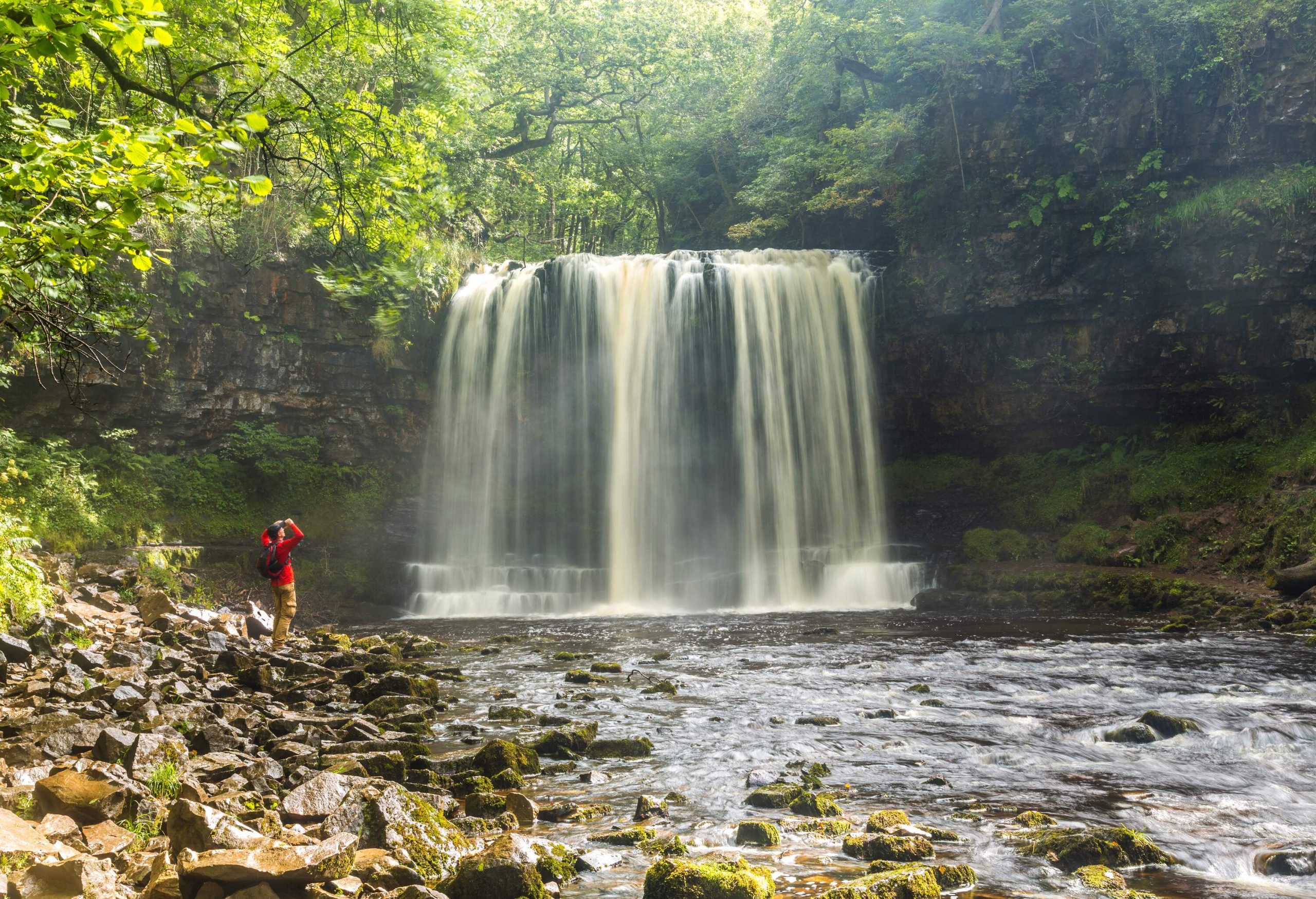 A tourist in awe of a stunning waterfall descends on a rocky path in a forest of lush green foliage.