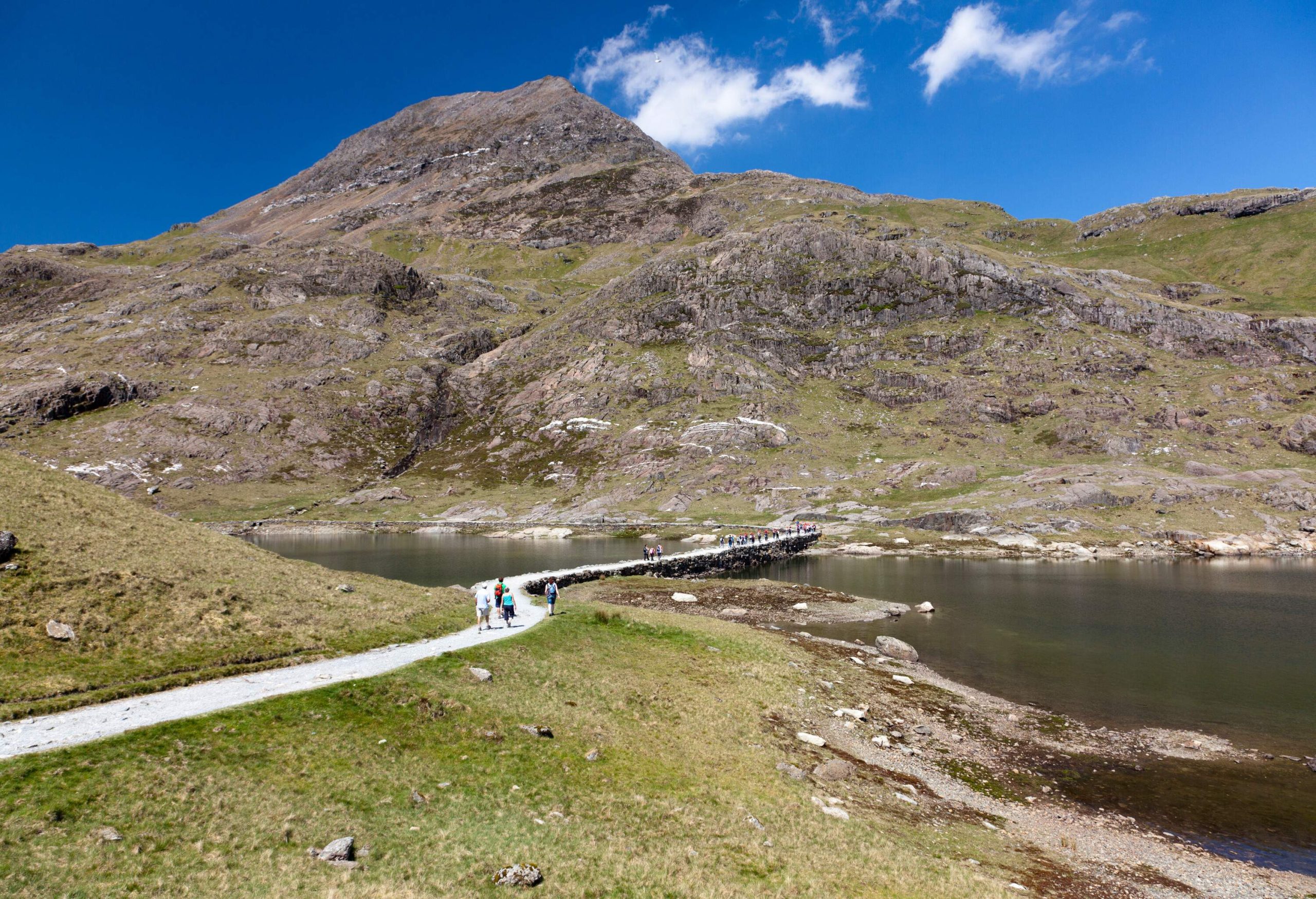 People walking across a footbridge over a lake flowing through the mountains.