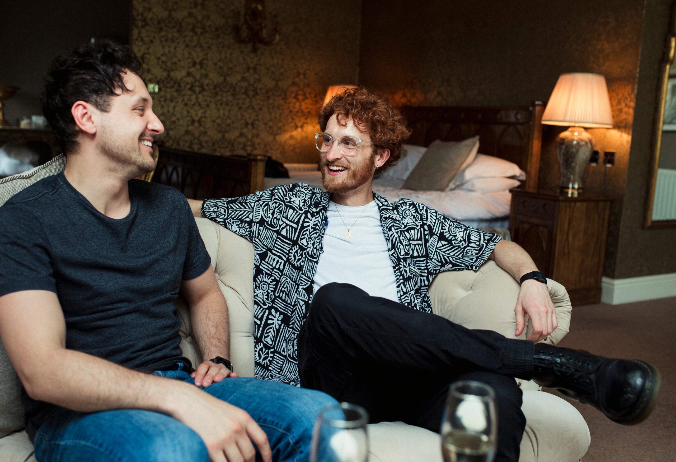 Two male friends sitting on the sofa share stories happily in their hotel room.