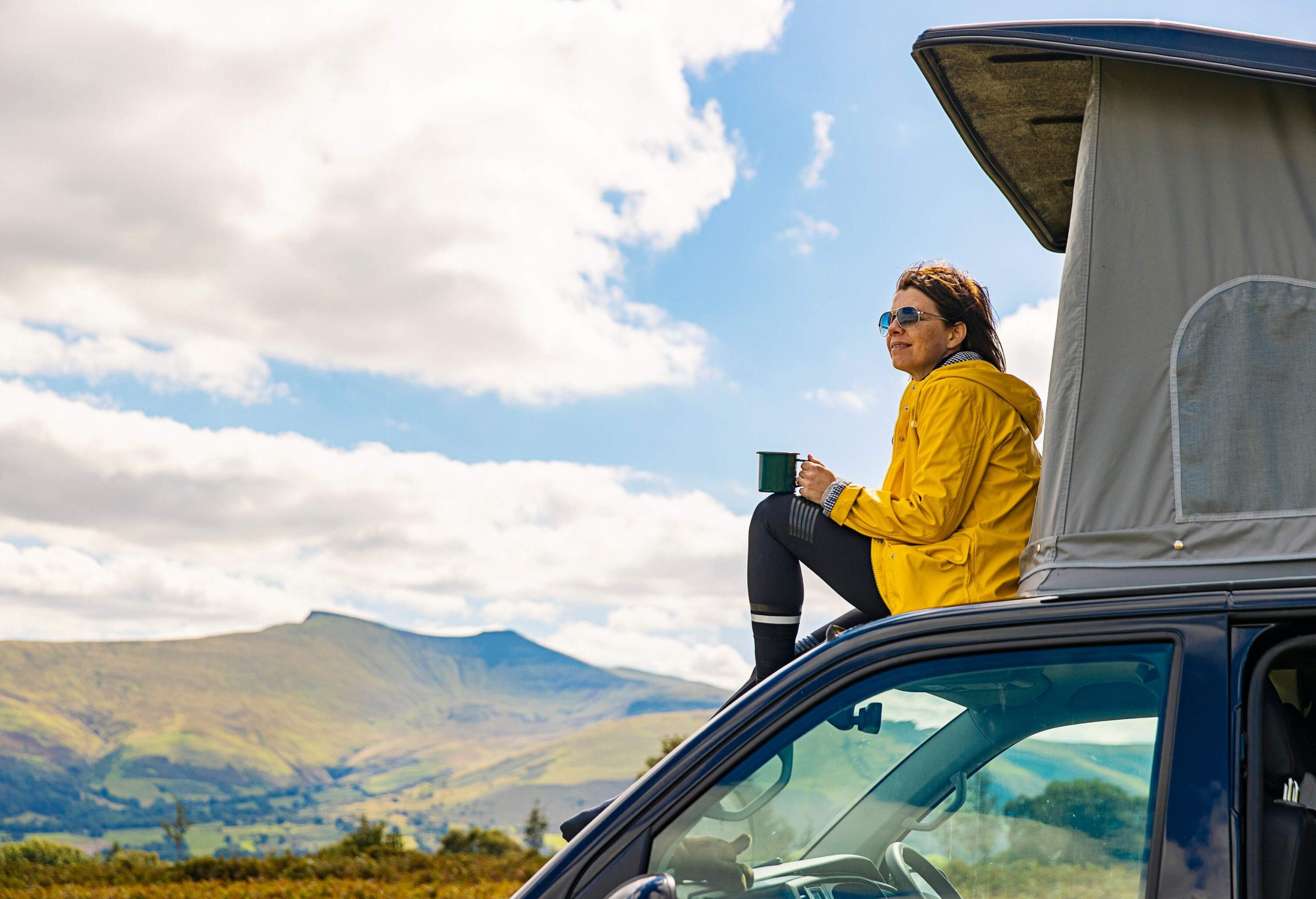 A woman in a yellow jacket enjoys a cup of coffee as she sits beside a tent on top of her car.