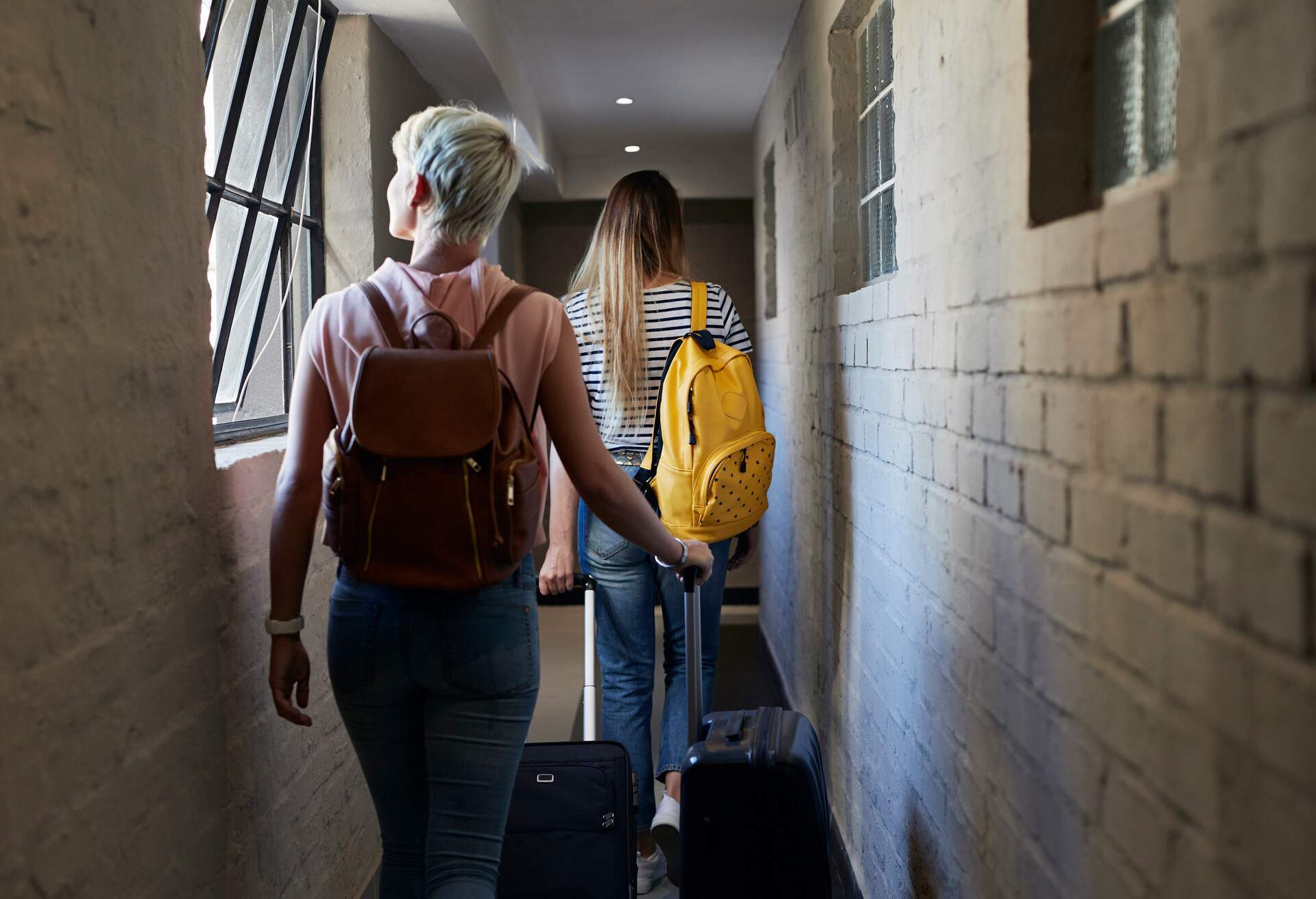 Two female friends walk into the hallway with their backpacks and luggage.