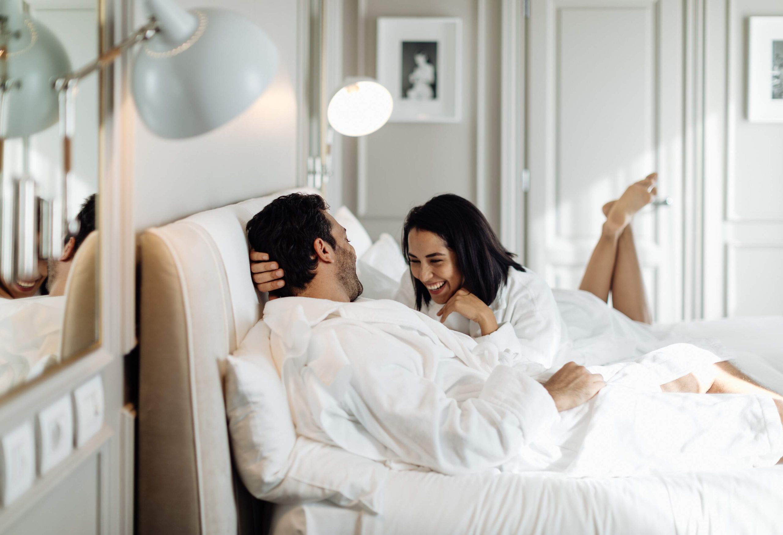 A couple laughing together in bed while wearing white plush robes.