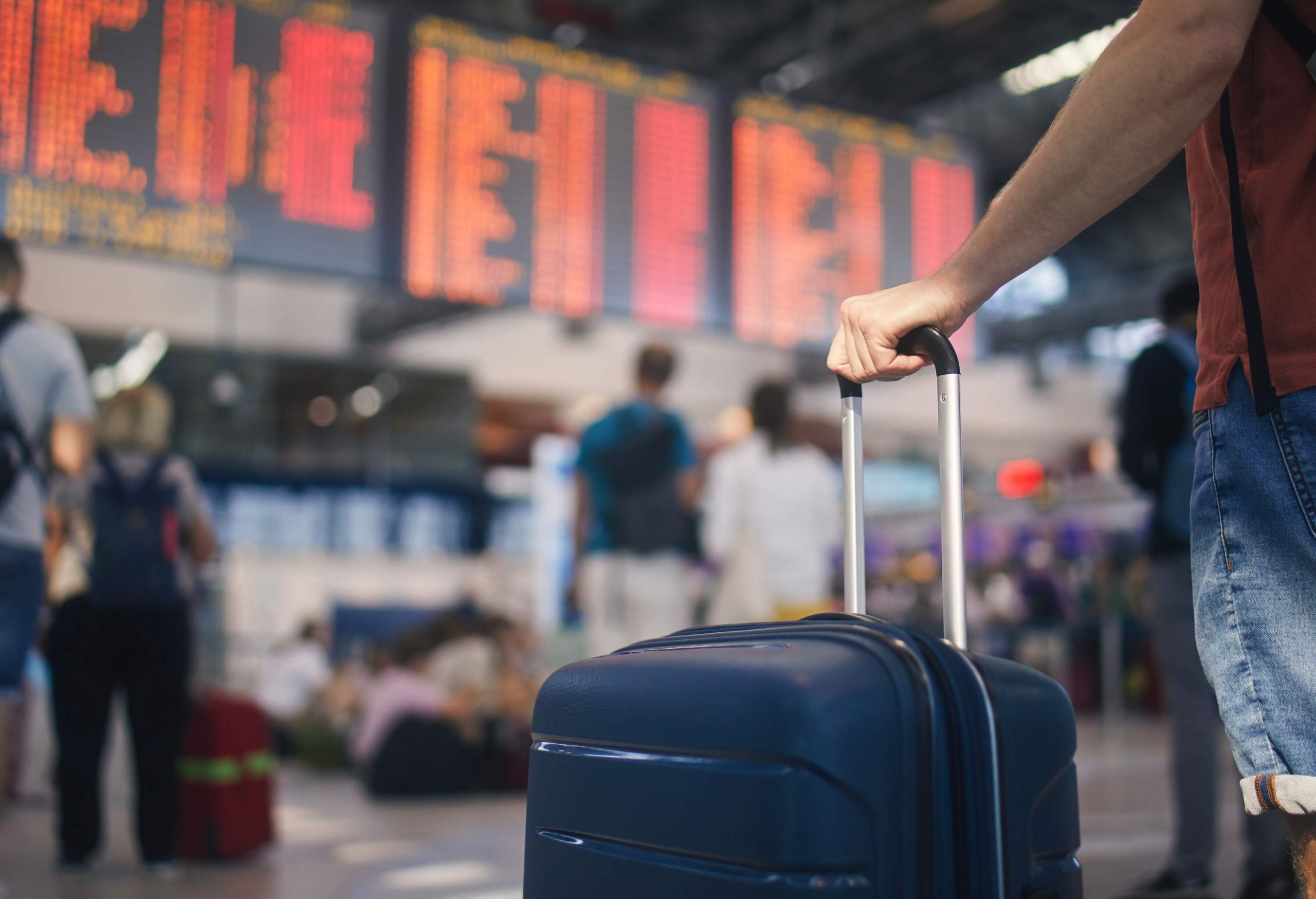 Traveling by airplane. Man waiting in airport terminal. Selective focus on hand holding suitcase against arrival and departure board. Passenger is ready for travel.