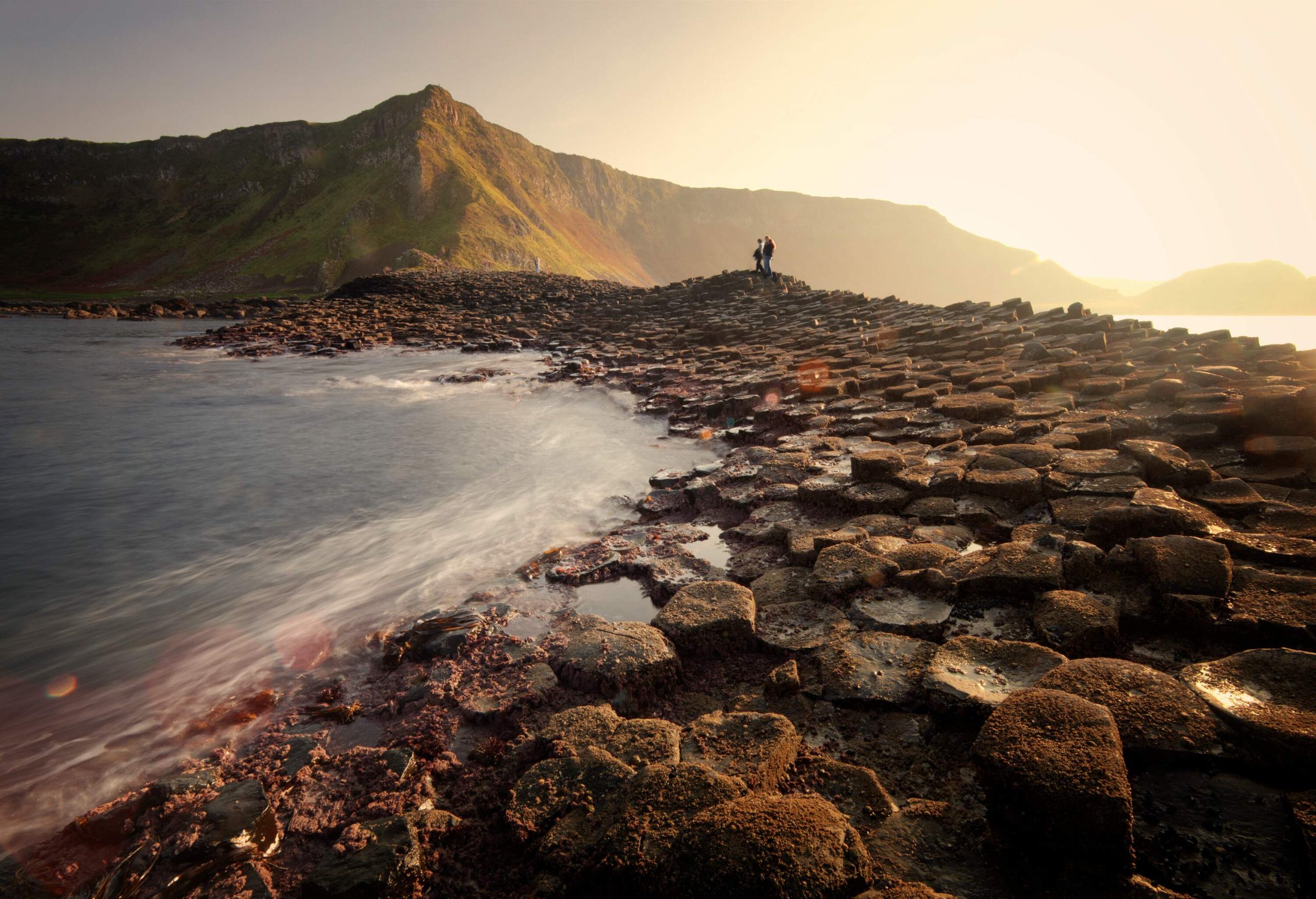 Two people stand on the interlocking columns along the shore with a background of cliffs at sunset.