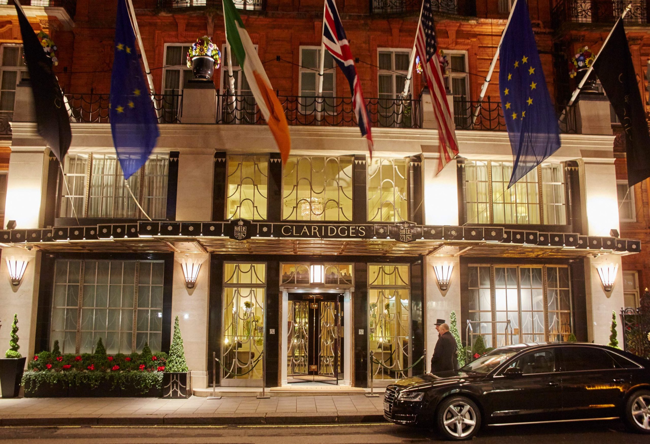 A black car parked outside a hotel with glass façade with a variety of world flags hanging on the balcony.