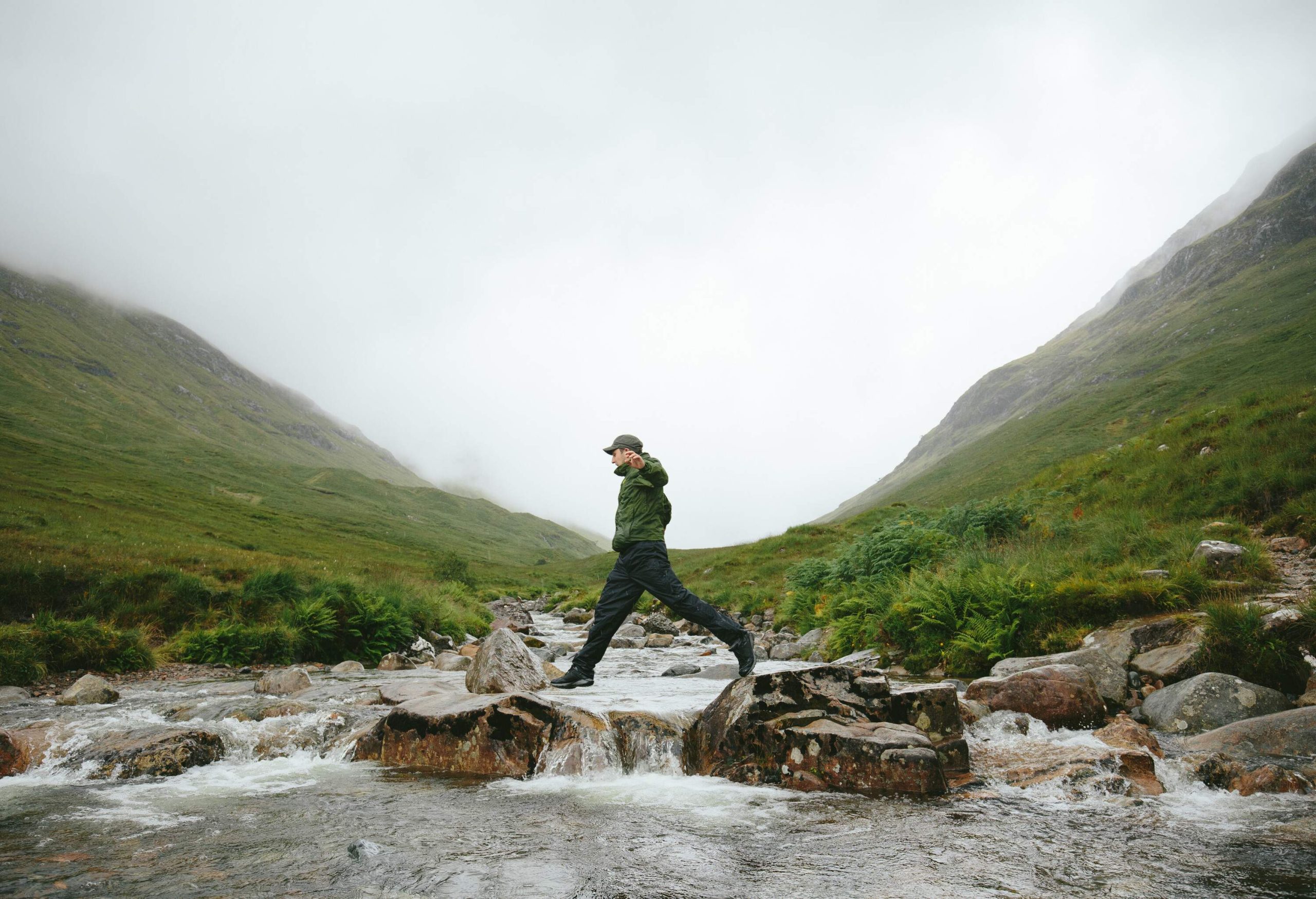 A man skillfully balances on the rocks within a gushing river, surrounded by the awe-inspiring beauty of lush mountains.