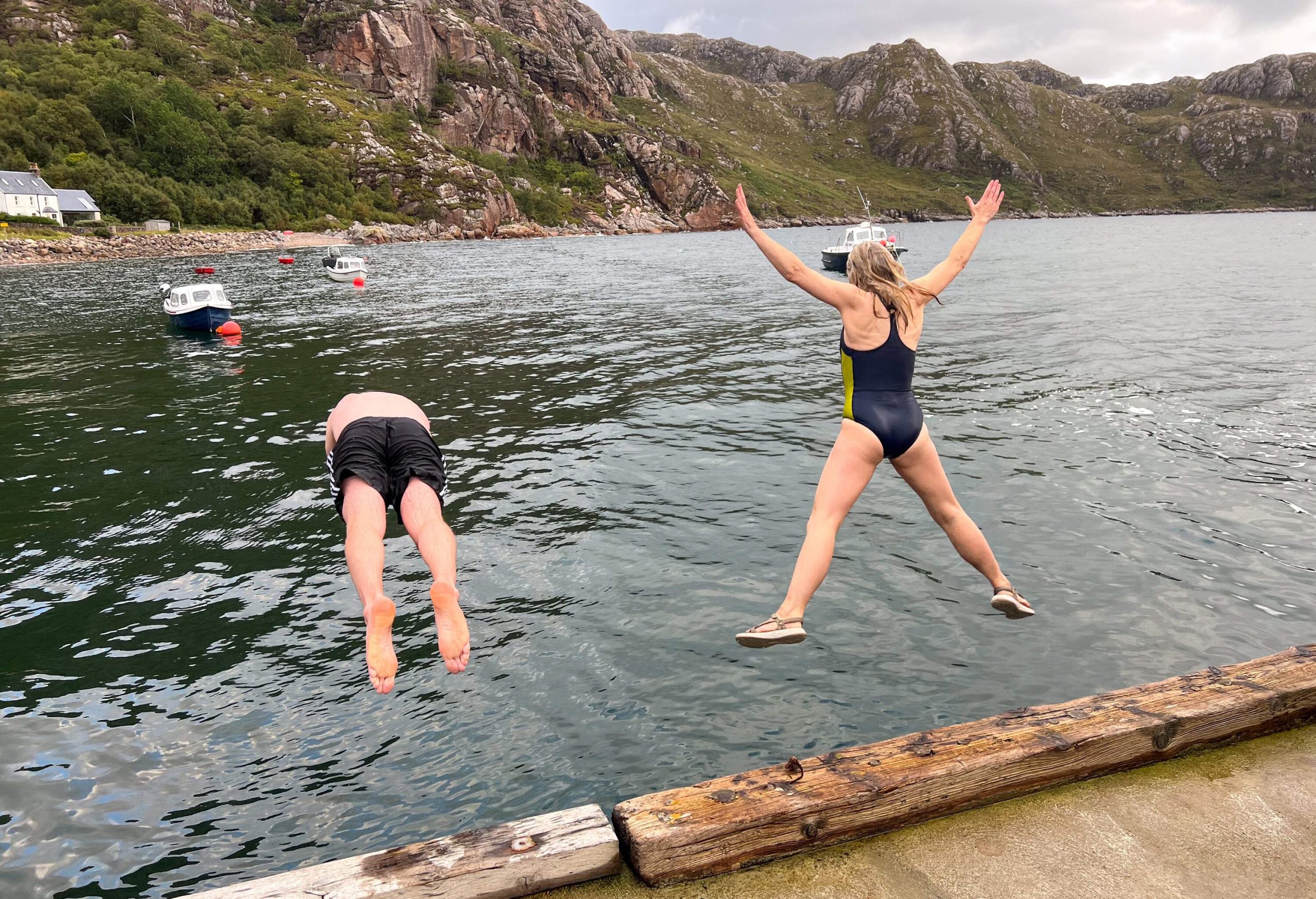 A full shot of two unrecognisable friends mid-air jumping into a loch at the same time on the side of Loch Torridon near the village of Diabaig in the west highlands of Scotland, wearing swimming gear.