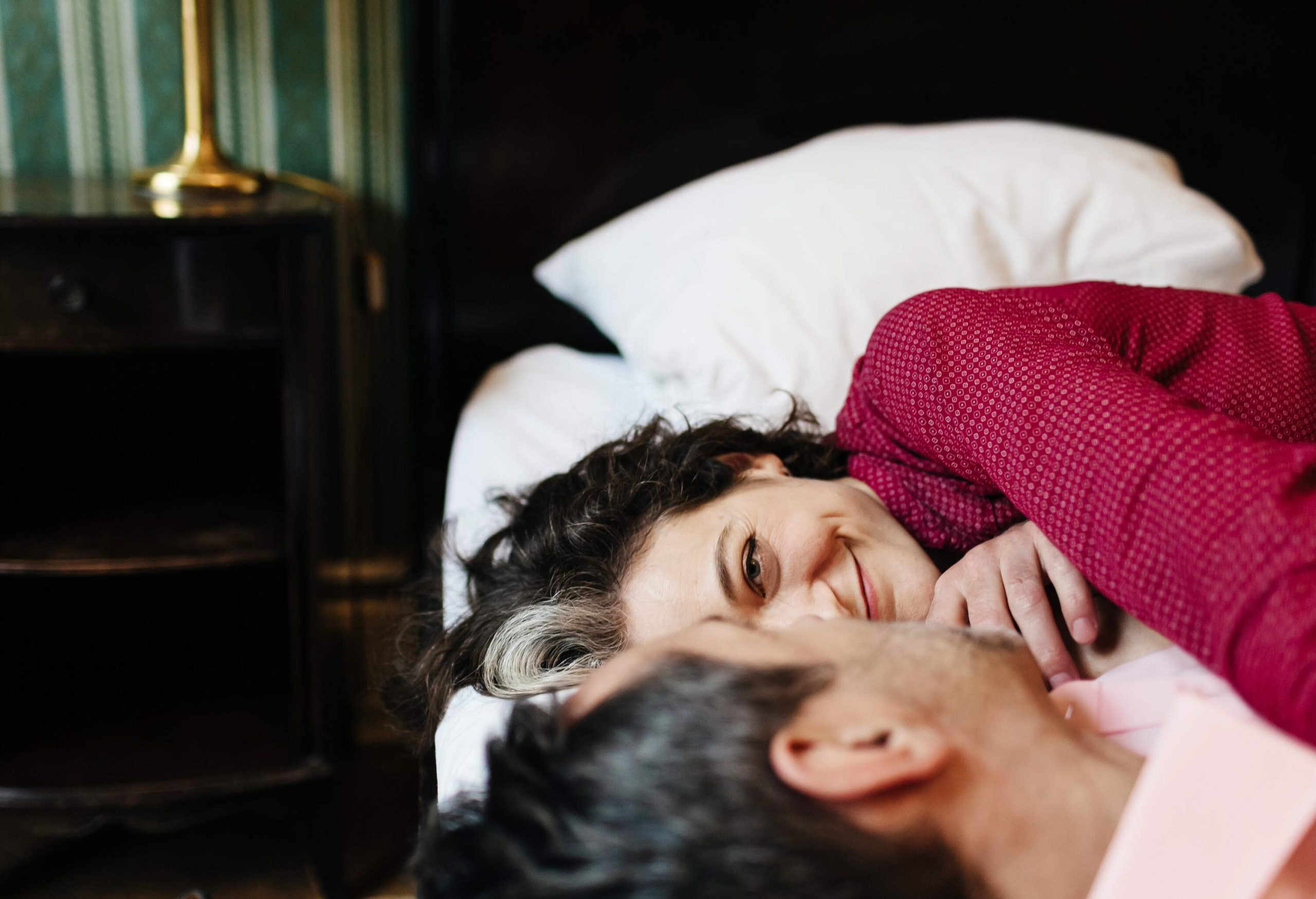 A middle-aged woman smiles gladly at her partner as they lie on the bed.