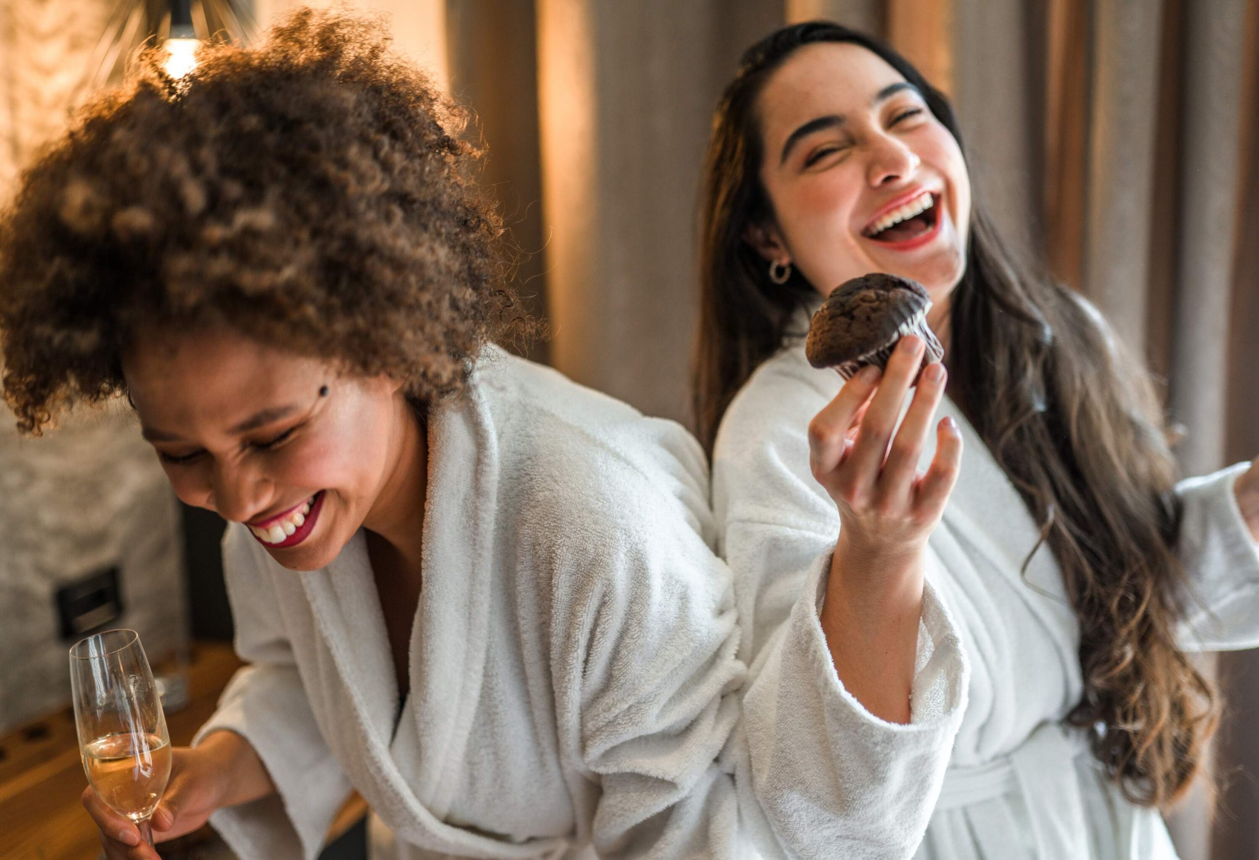 Two young women of mixed race share a playful moment in a hotel room, donning bathrobes and holding glasses of wine, with one of them also clutching a delicious chocolate muffin.