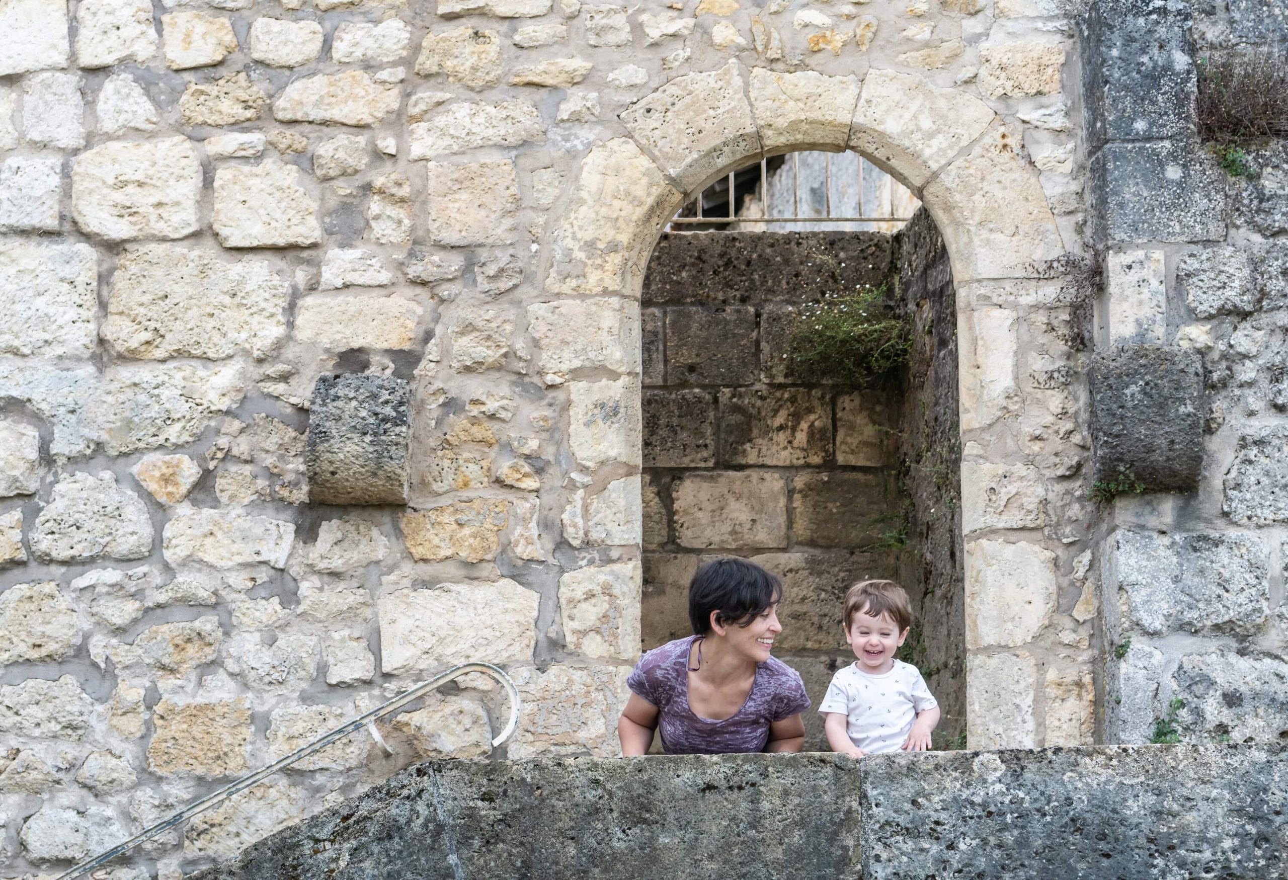 A mother looks at a boy smiling on a terrace under a limestone arch.