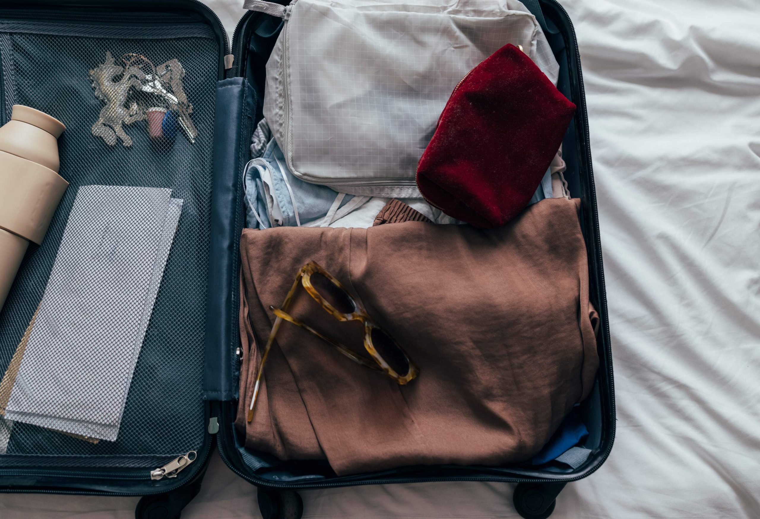 A cropped close-up of a grey spinner luggage from above on the bed on a holiday or a business trip containing sunglasses, a cosmetic bag, a water bottle, keys, clothes and plane tickets.