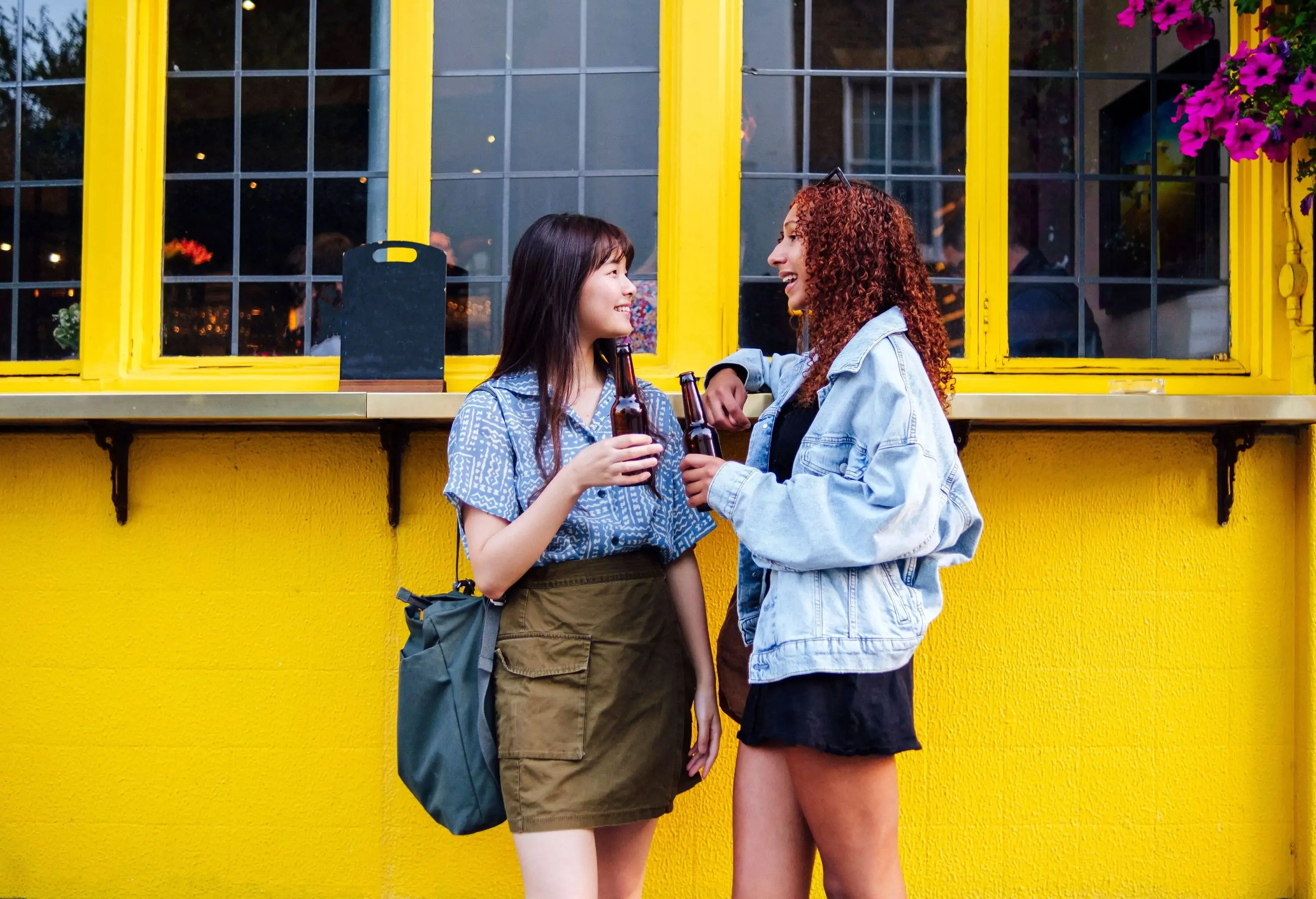 Two female friends chit-chat while holding a bottle of drinks, standing against the yellow wall outdoors.