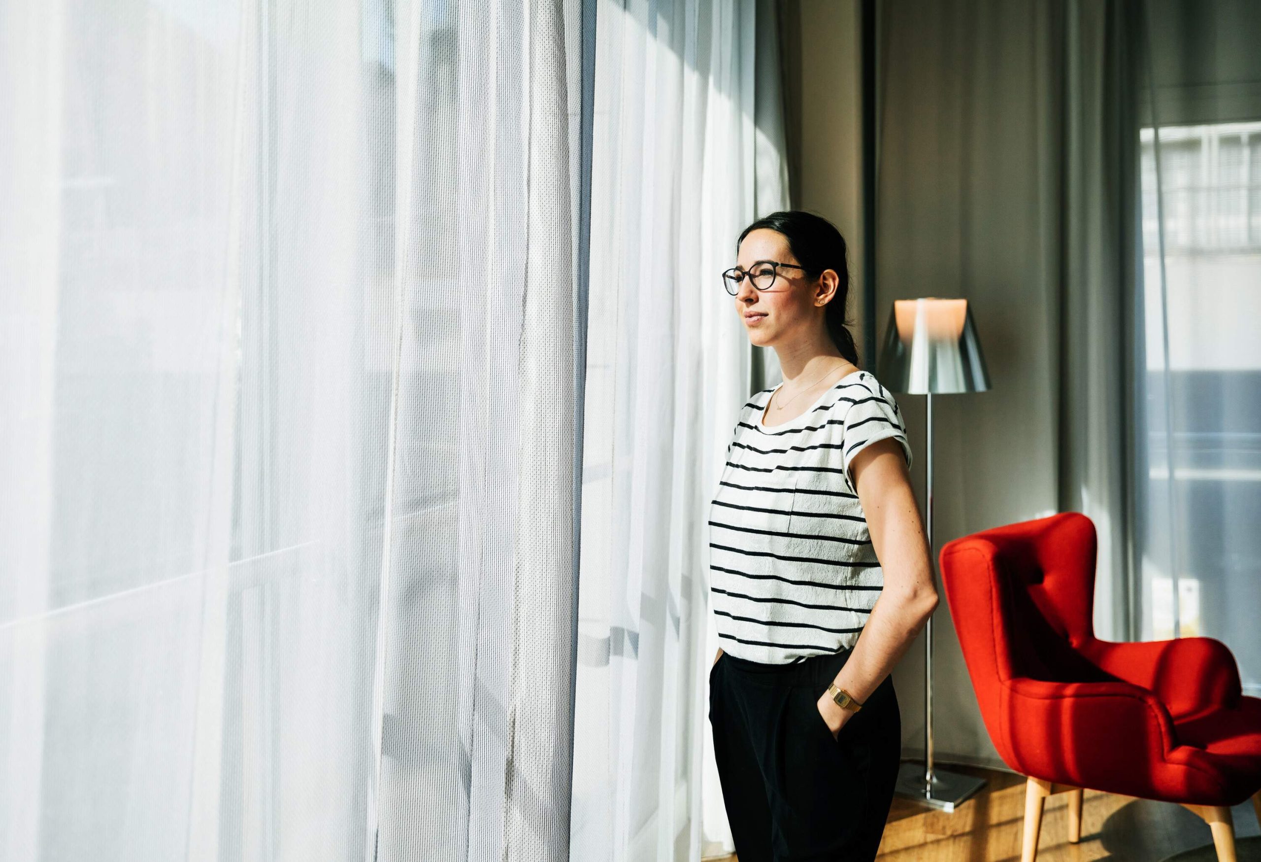 A woman in a black and white striped top standing by the window with her hands in her pant pockets.