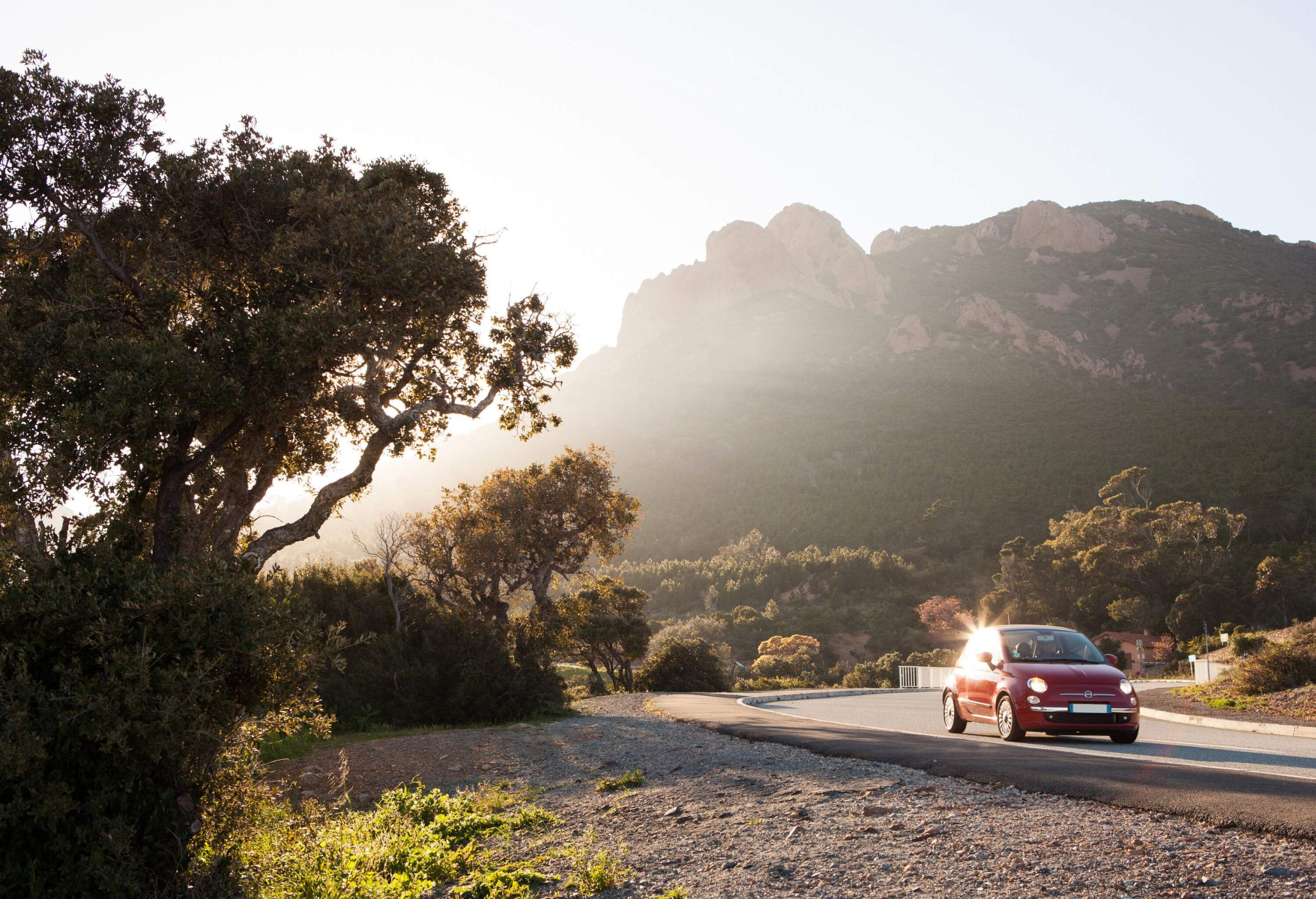 A small red car transverses a curved road with a view of a rugged, steep mountain behind.