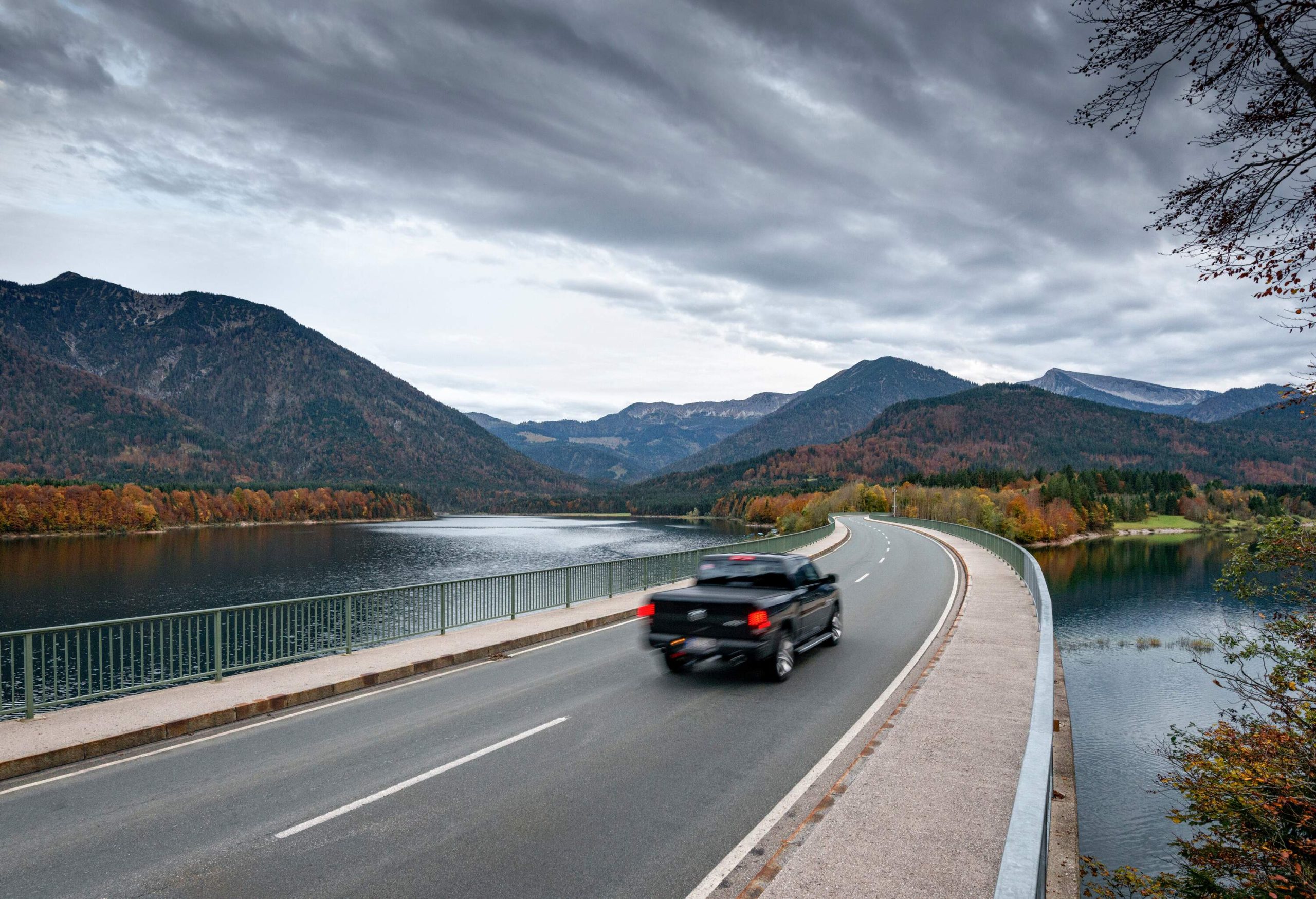 A pickup truck drives along a winding road next to a lake in the forest-clad mountains in the background.