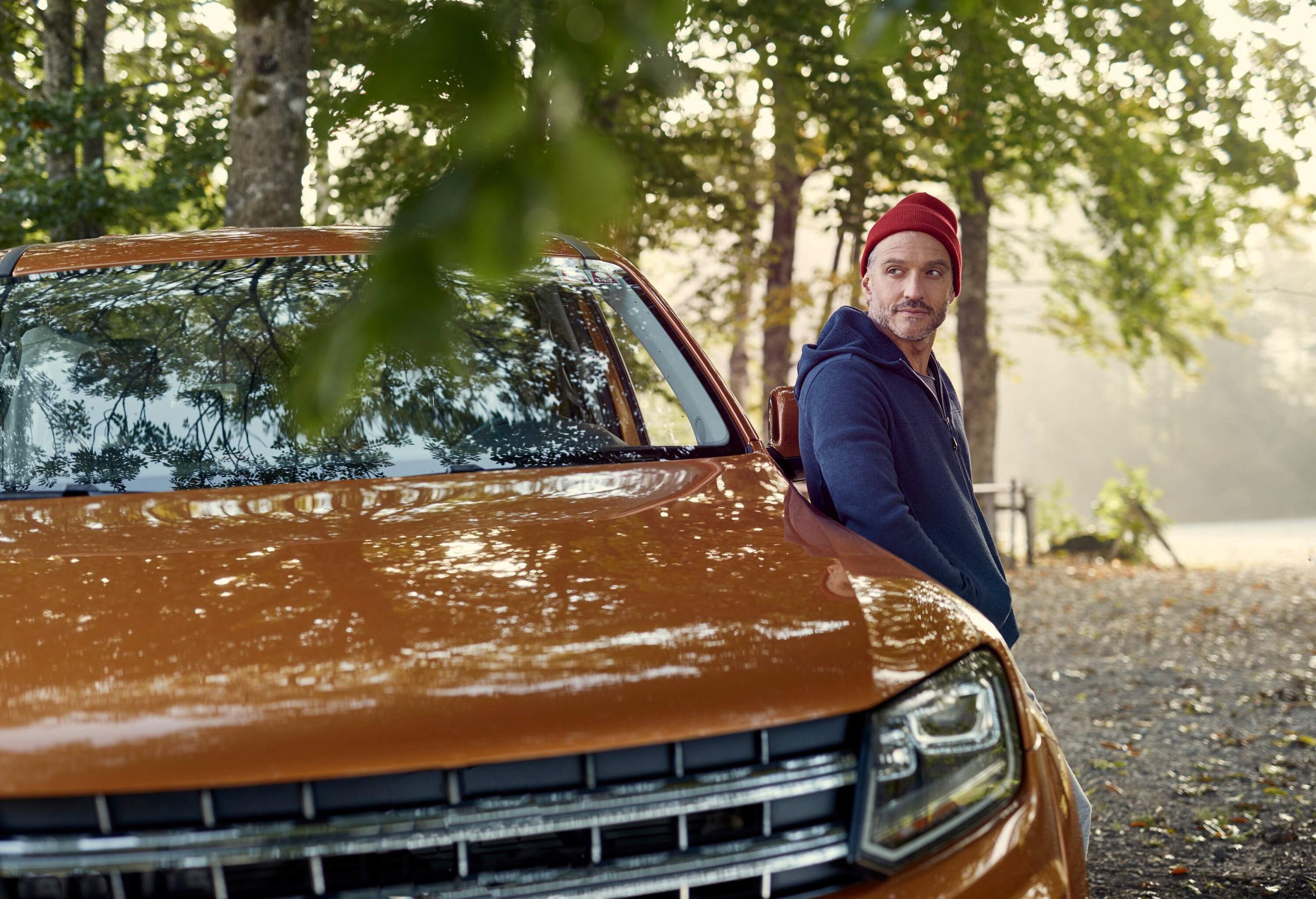 Man glances back while leaning on a brown pickup truck wearing a maroon bonnet and a navy hoodie.