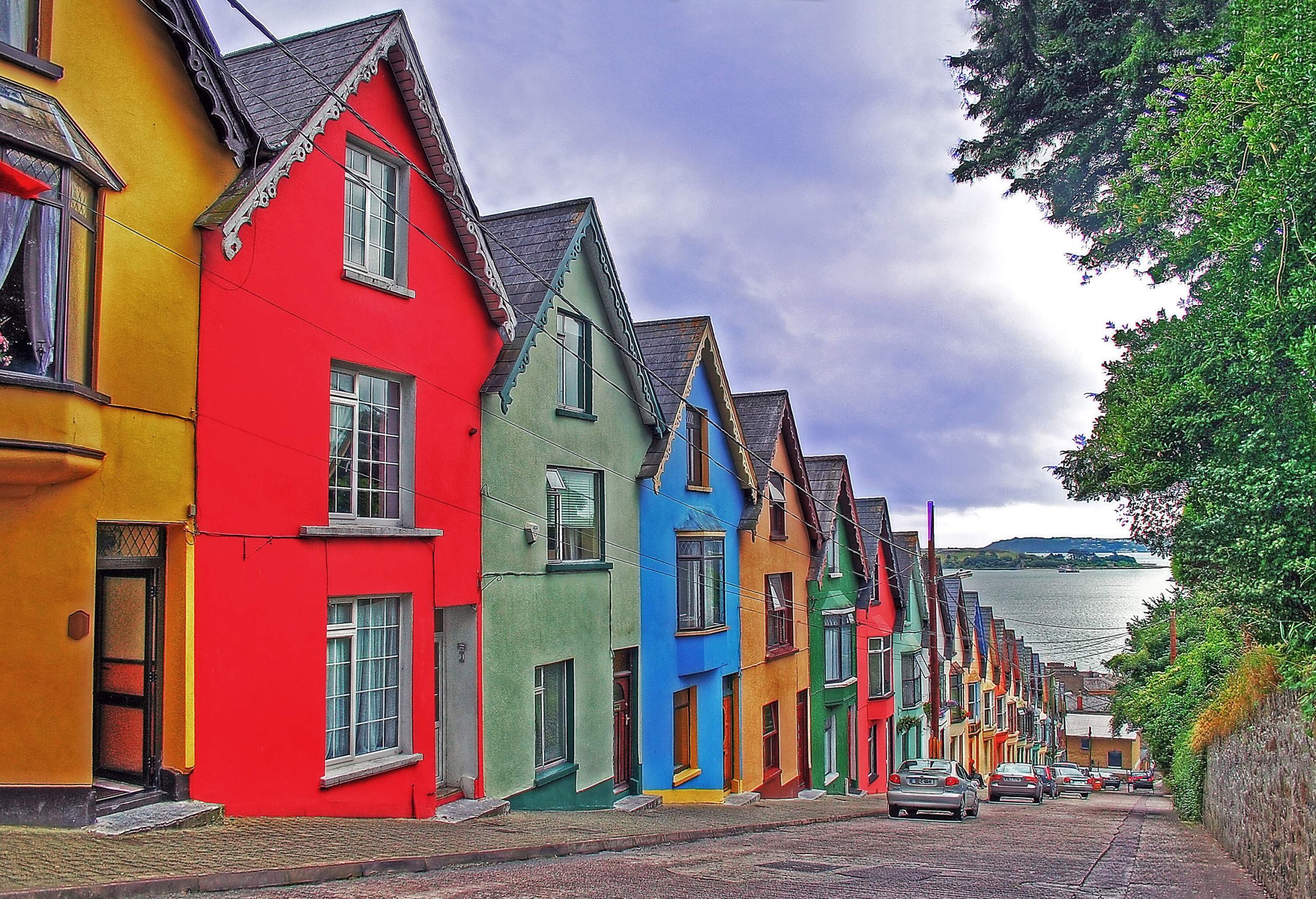 A row of colourful terraced houses along a downhill street with parked cars.