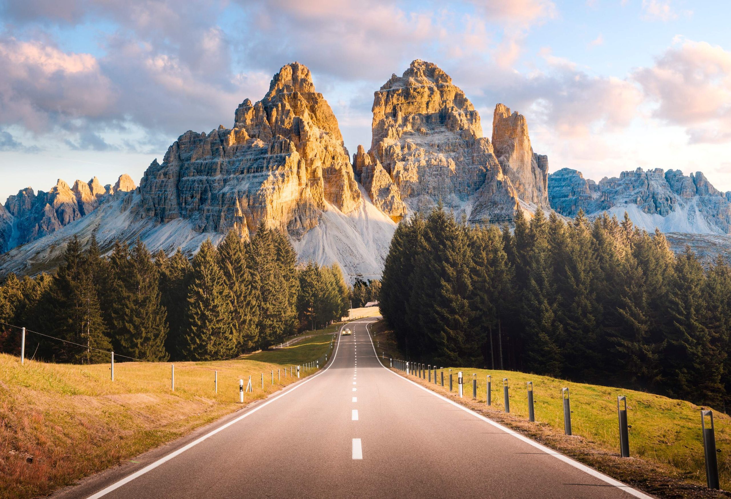 A car driving down a road that runs through coniferous trees and along the foothills of the Tre Cime di Lavaredo mountains.
