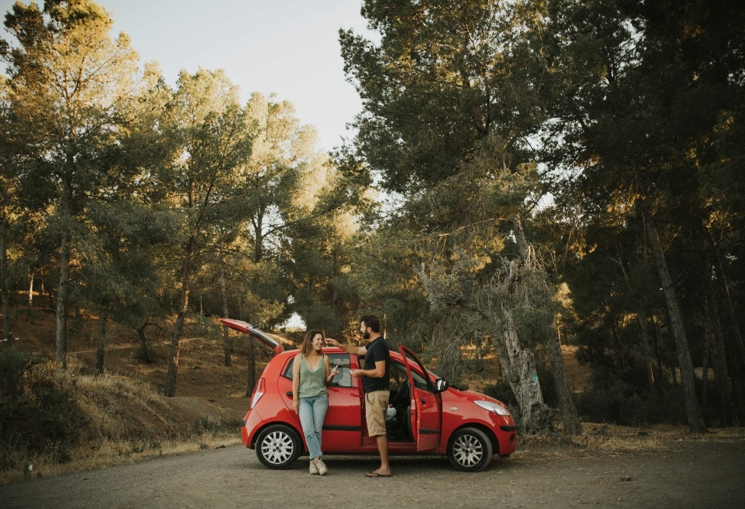 Two people standing on the side of a parked red car with open doors with a view of tall trees in the back.