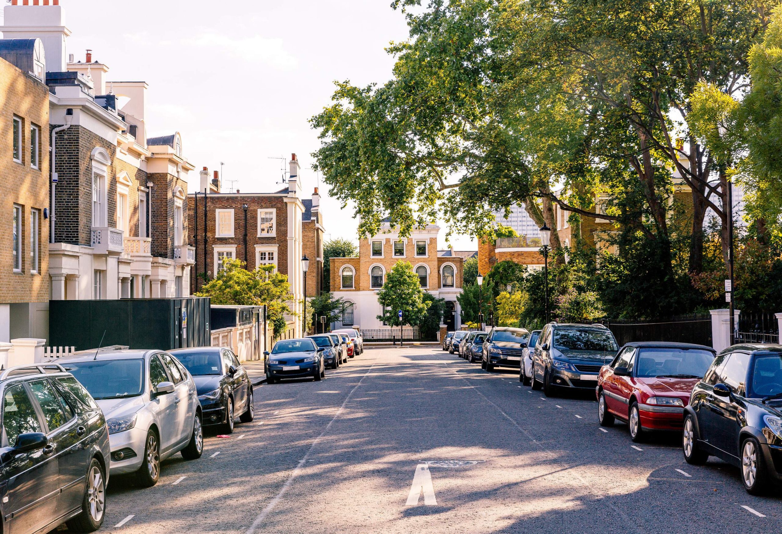 A paved road lined with parked cars on the sidewalk and beautiful classic townhouses with brick facades.