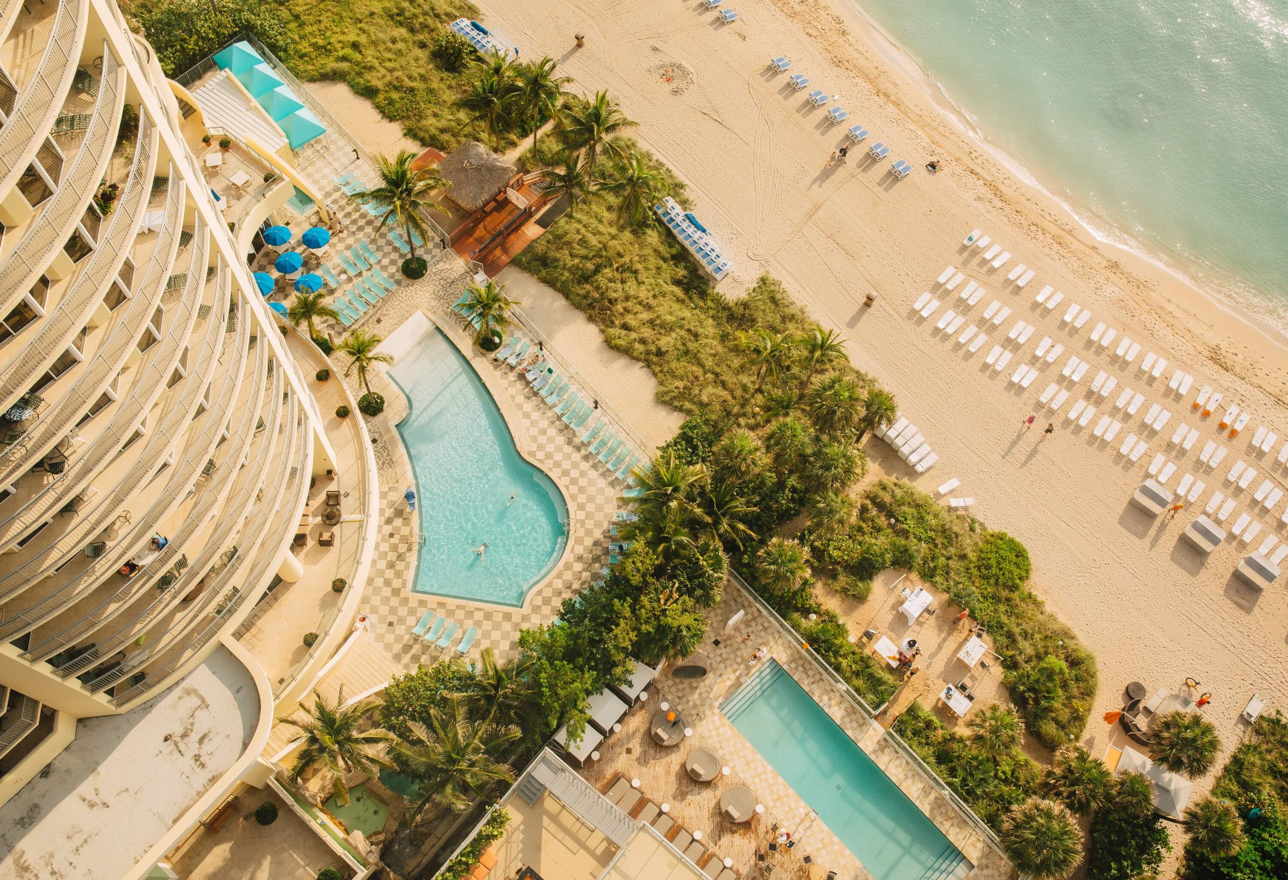 An aerial view of a resort with tall residential buildings overlooking the swimming pools facing the beach.