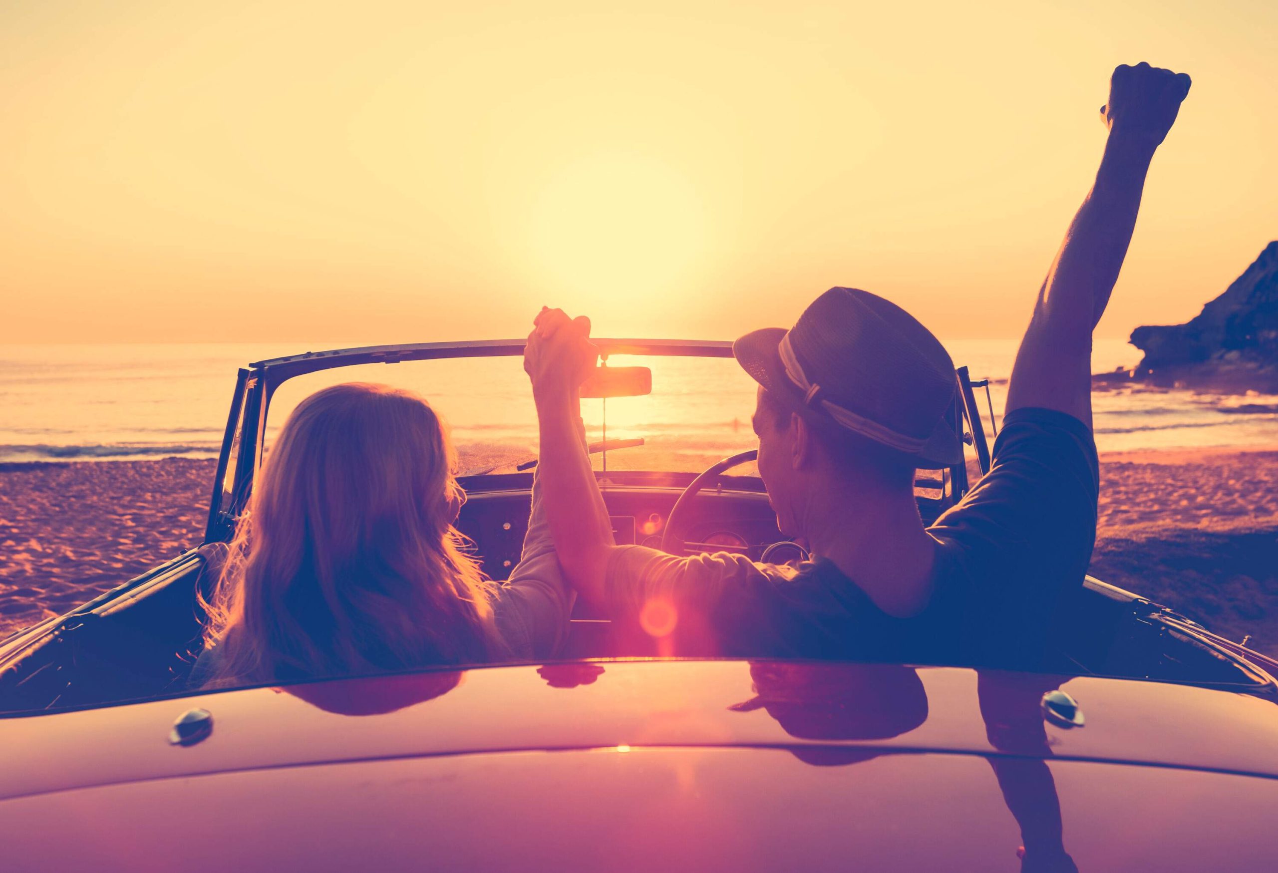 Rear view of a happy couple in a convertible car parked on the beach, holding hands, raising their hands while watching the scenic sunset.