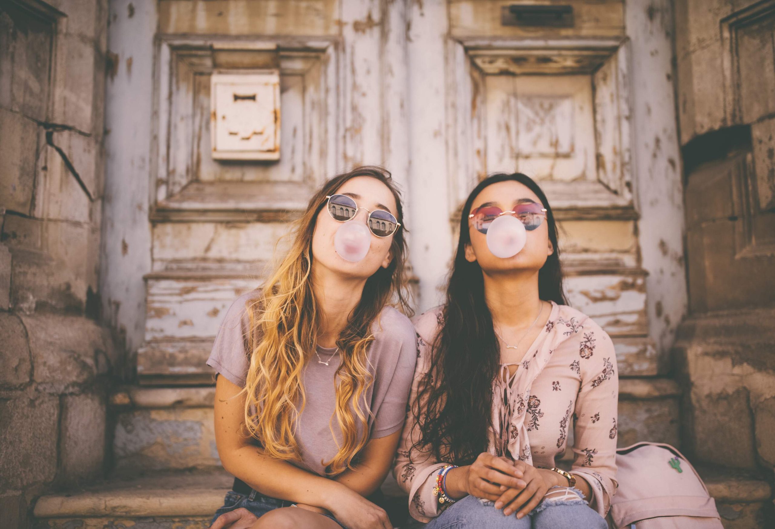 Two women in casual clothes and sunglasses sit beside each other as they blow bubbles with gum.
