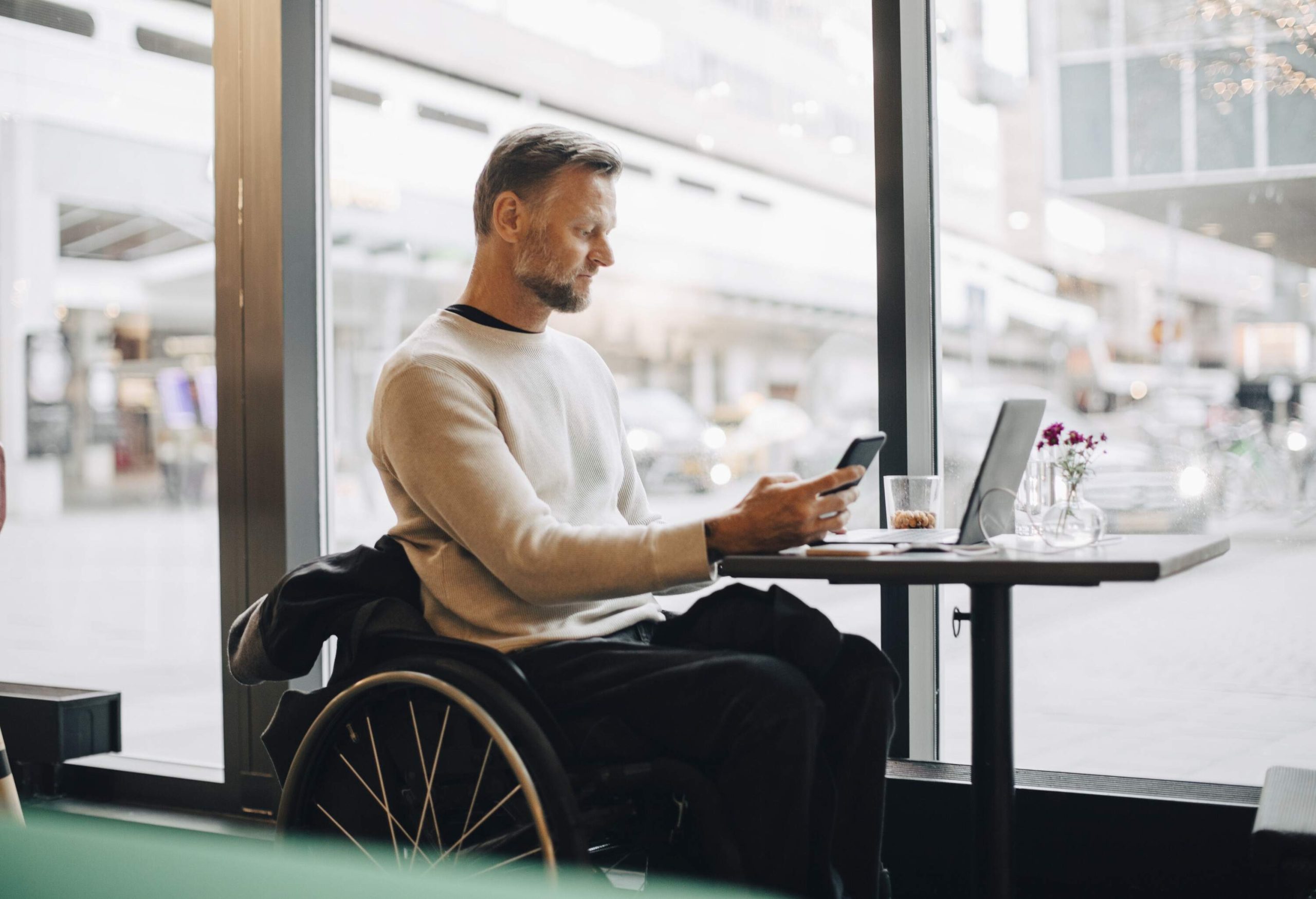 A man in a wheelchair looks at his mobile phone with his laptop on a table in a restaurant.